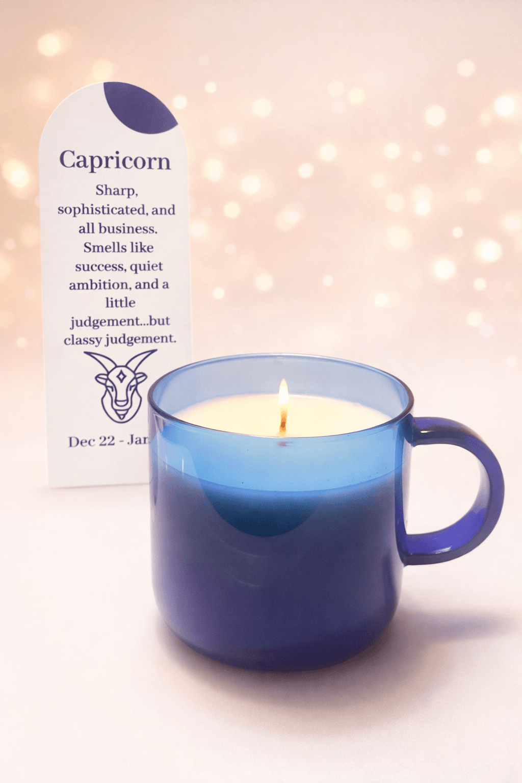 Blue candle in a mug with a zodiac sign card for Capricorn in the background, soft pink bokeh lights, and a white surface.
