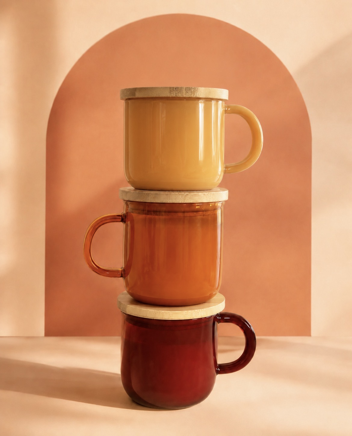 Three stacked ceramic mugs in yellow, orange, and red colors on a wooden surface with a pink arch background.