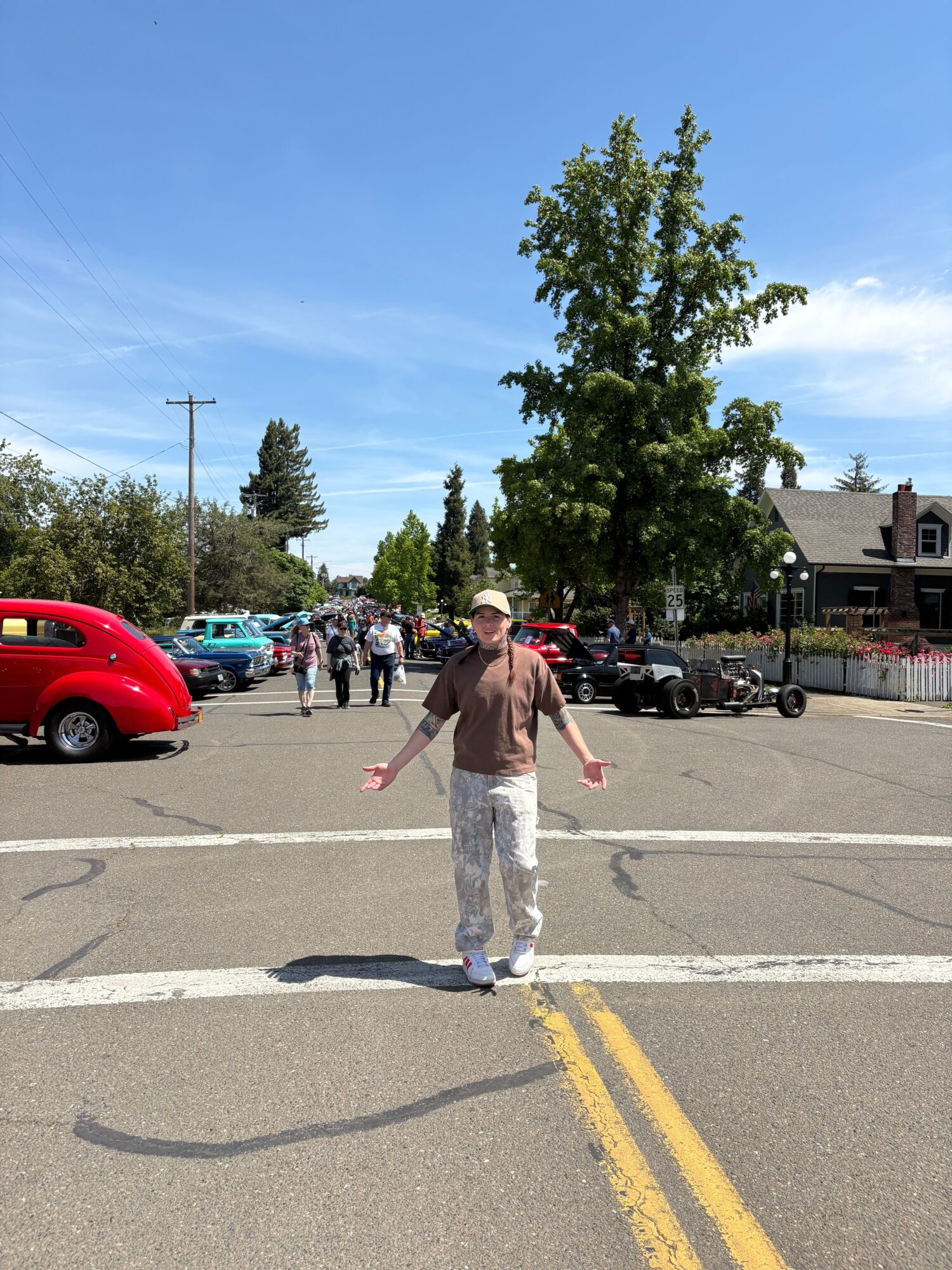 Person standing in a parking lot with trees and cars, waving with one hand, under a blue sky.