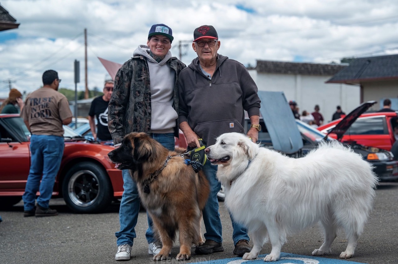 Two men stand with large dogs in front of classic cars at an outdoor event, cloudy sky overhead.