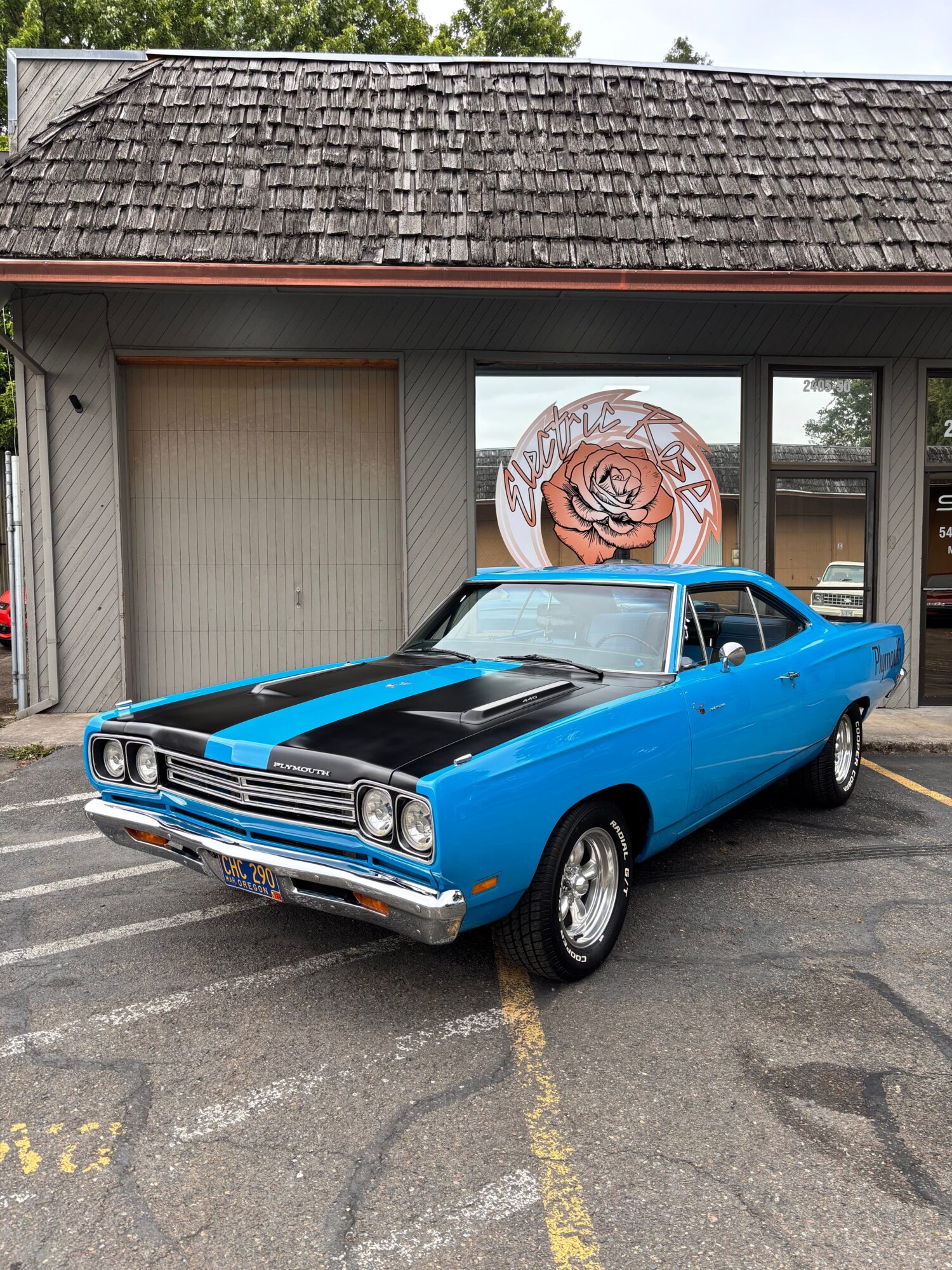 Blue vintage car parked in front of a building with a rose mural, on a paved lot.