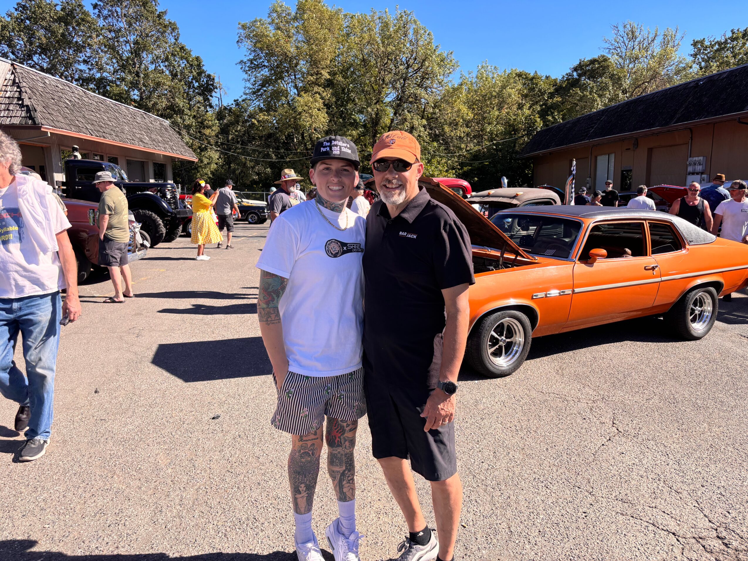 Two men stand outdoors near a vintage orange car, with other people and buildings in the background.
