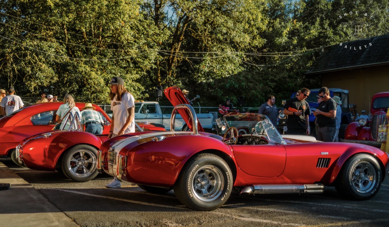 Red vintage sports cars parked outdoors with people around, trees in the background, and a building on the right.