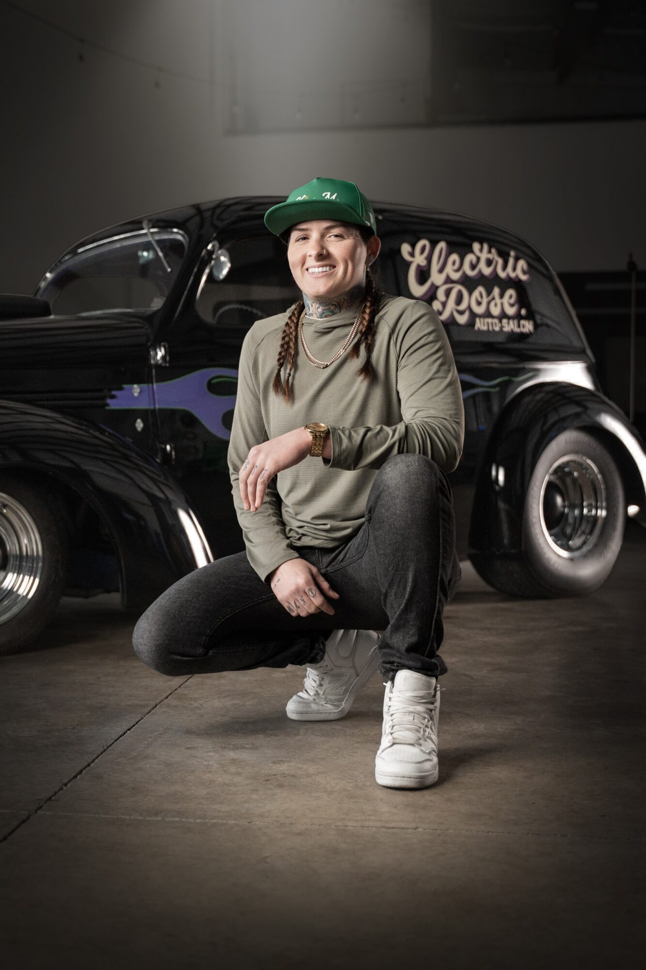 Person crouching in front of a vintage black car with a sign reading Electric Hope, smiling, wearing a green cap and casual clothing.