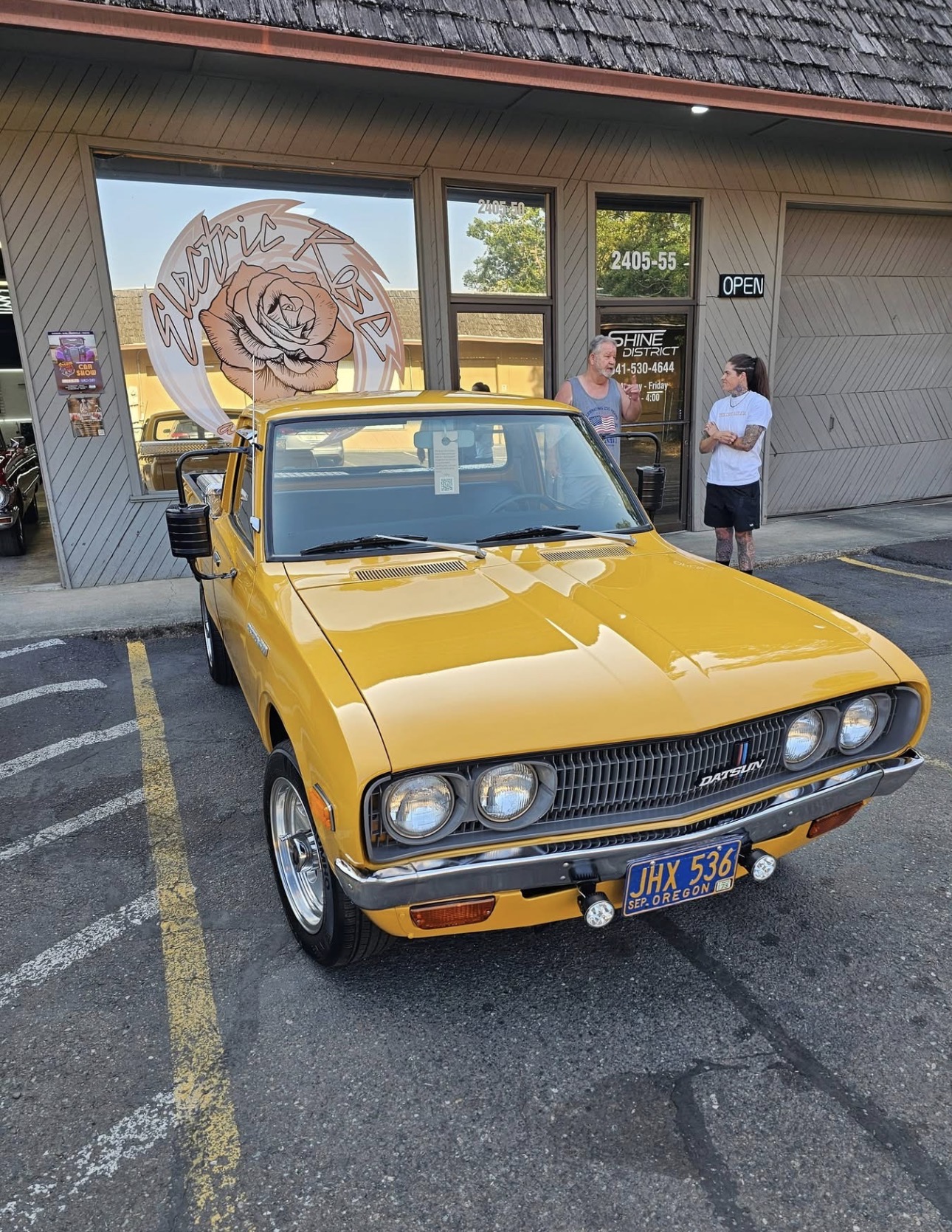 Yellow vintage car parked in front of a store with a large rose logo on the window, two people standing nearby.
