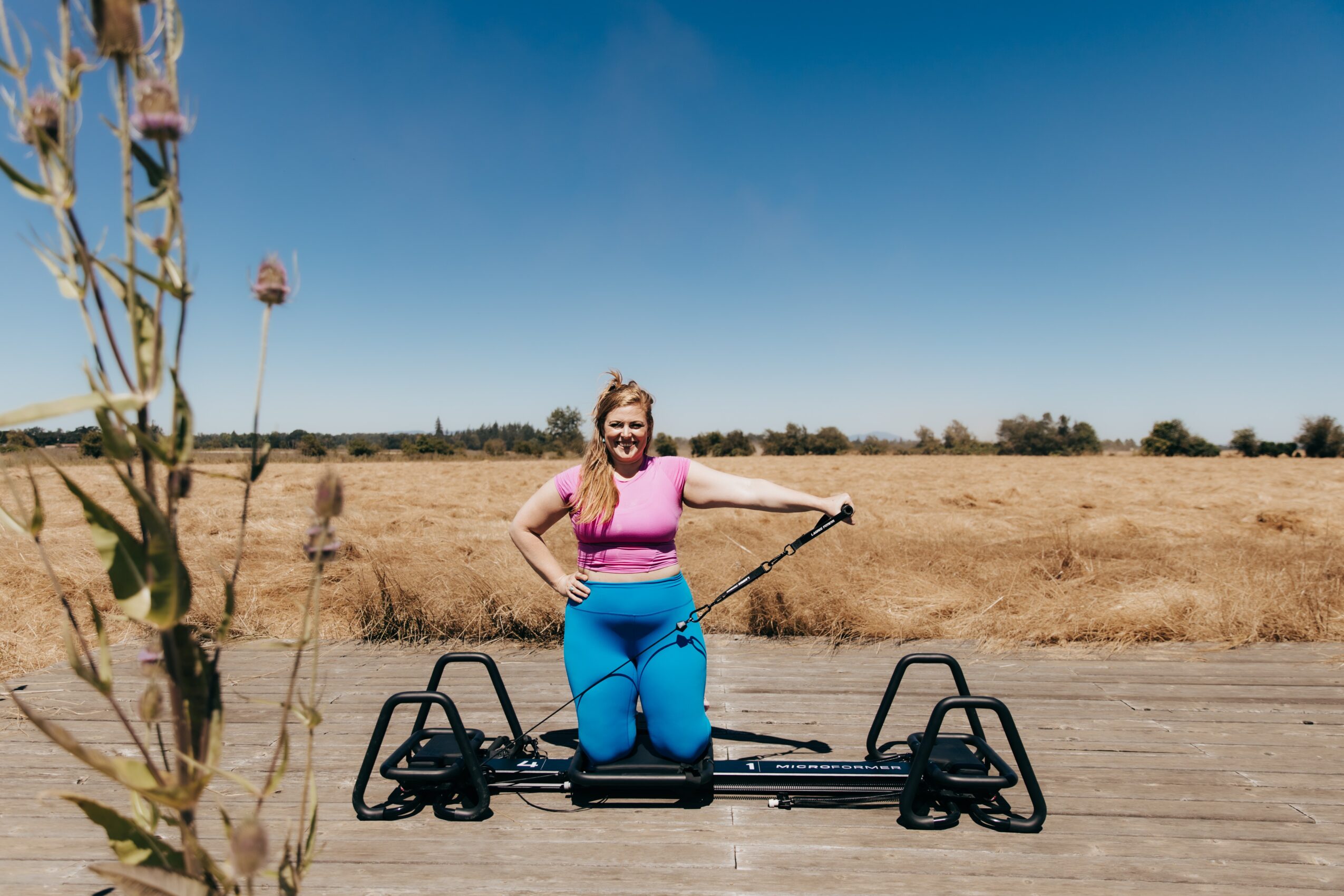 Woman sitting on a skateboard on a wooden path in a field, smiling, holding a stick, with a clear blue sky.