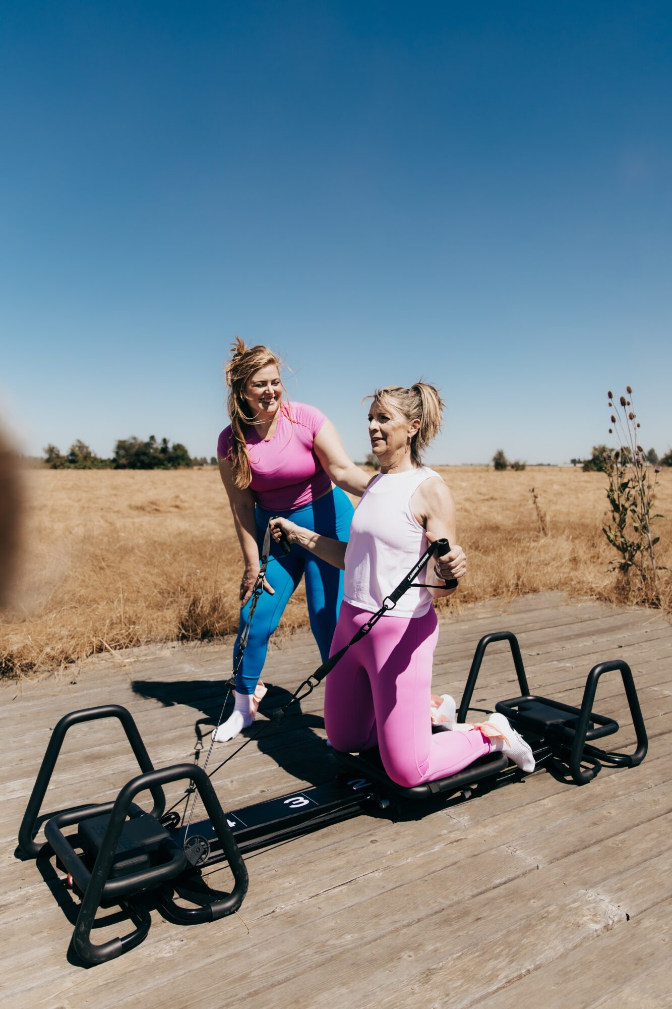 Two women outdoors, one kneeling on a fitness platform, the other standing and smiling, in a field under a clear blue sky.