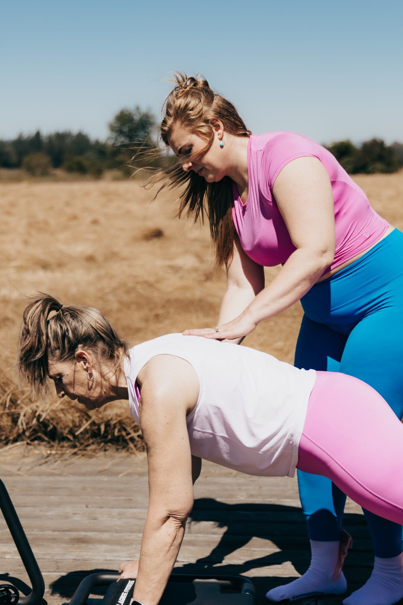Two women exercising outdoors on a sunny day, one in a plank position, the other assisting. Field and trees in background.