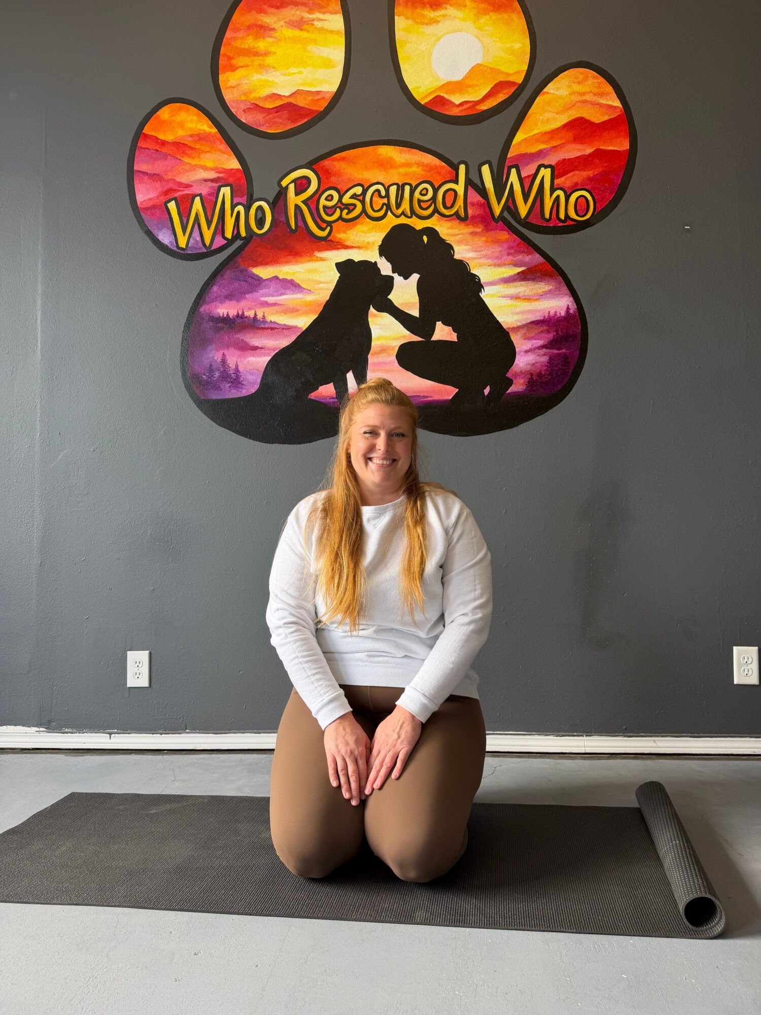 Person sitting on a gray mat in front of colorful wall art with a silhouette and text, indoors.