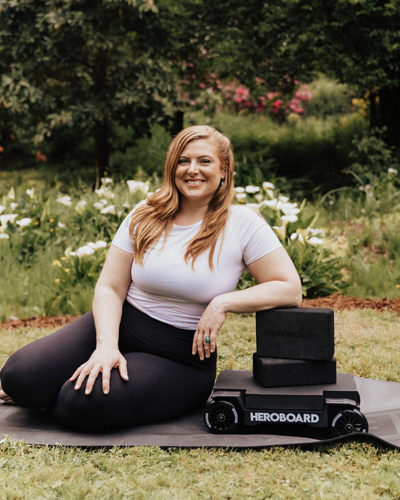 Woman sitting outdoors on a mat with a robot, smiling, surrounded by trees and flowers.