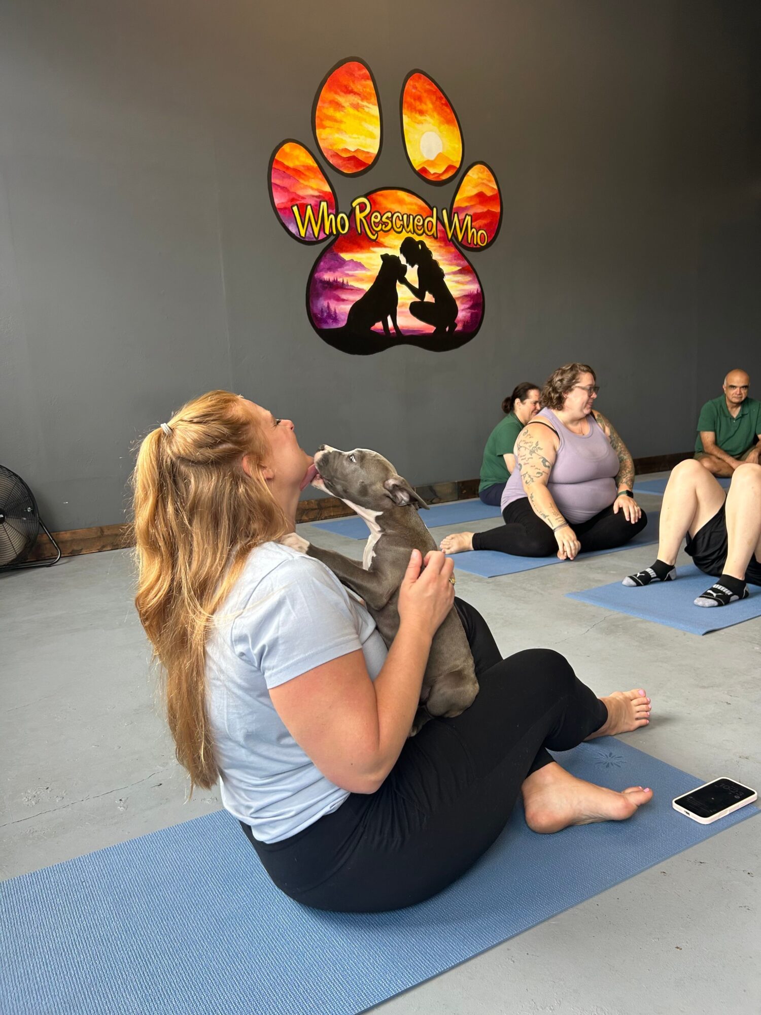 A woman with blonde hair sitting on a blue mat, holding a dog, with three children sitting nearby on the floor.