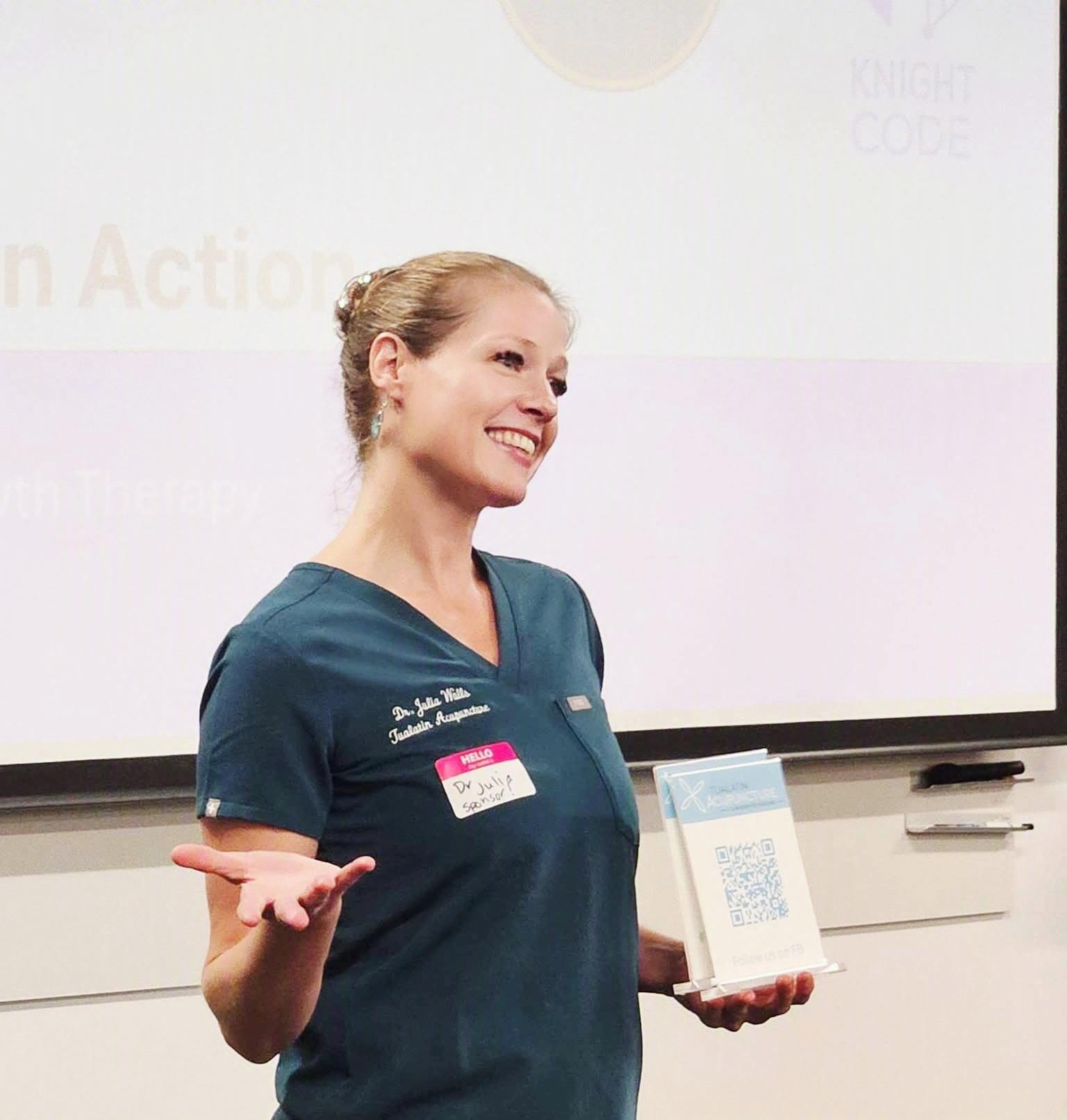Smiling woman in scrubs holding a plaque, gesturing with her hand in a classroom or conference room.