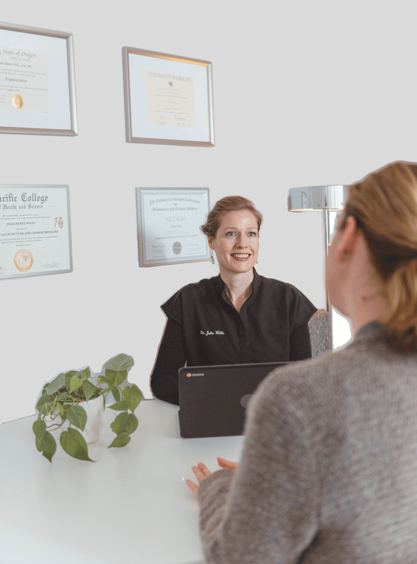 A woman with glasses and a gray sweater sits at a desk talking to a smiling woman in a black uniform, with certificates on the wall behind them.