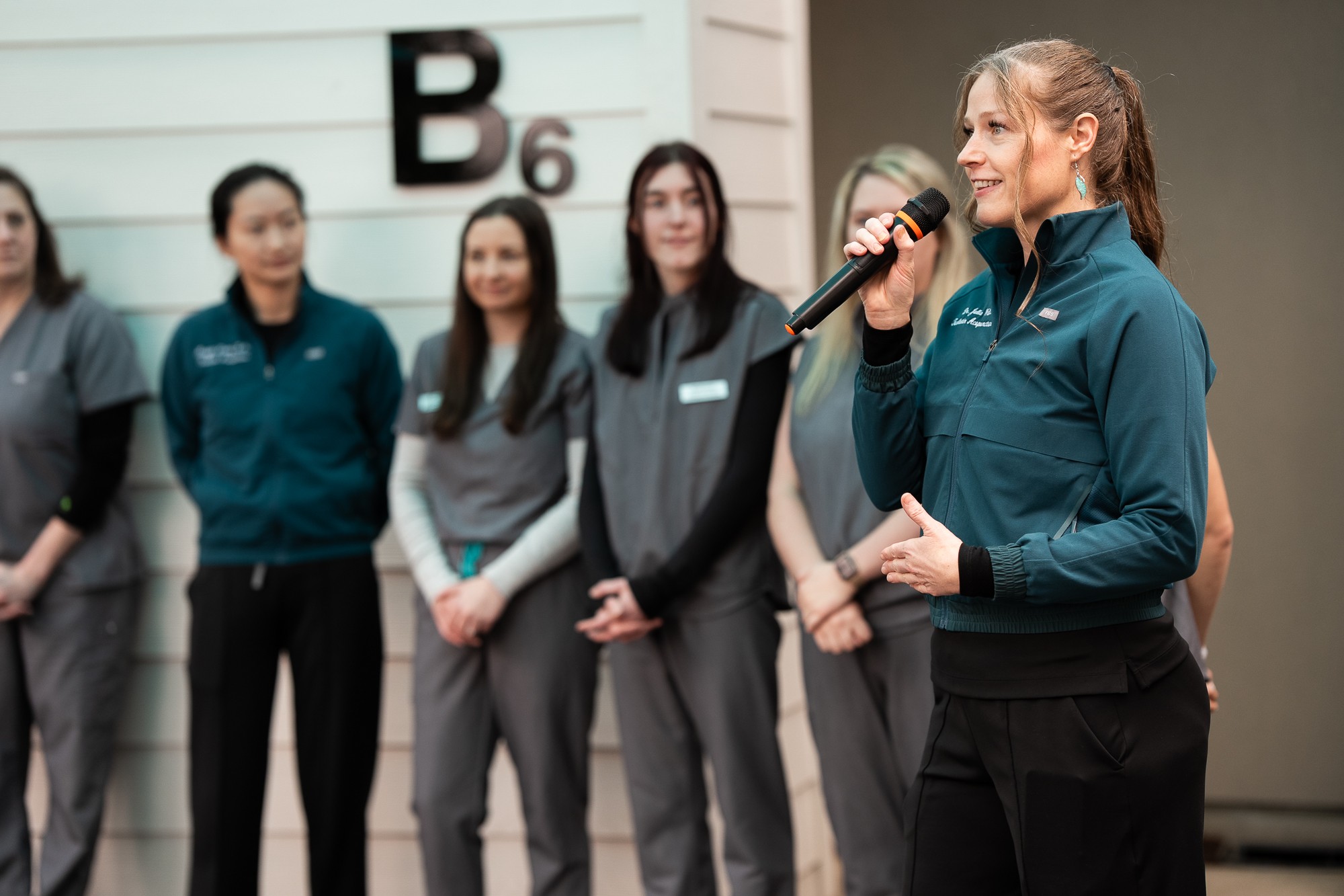Woman speaking into microphone in front of group of women, indoors, B6 sign visible, smiling, wearing teal jacket.