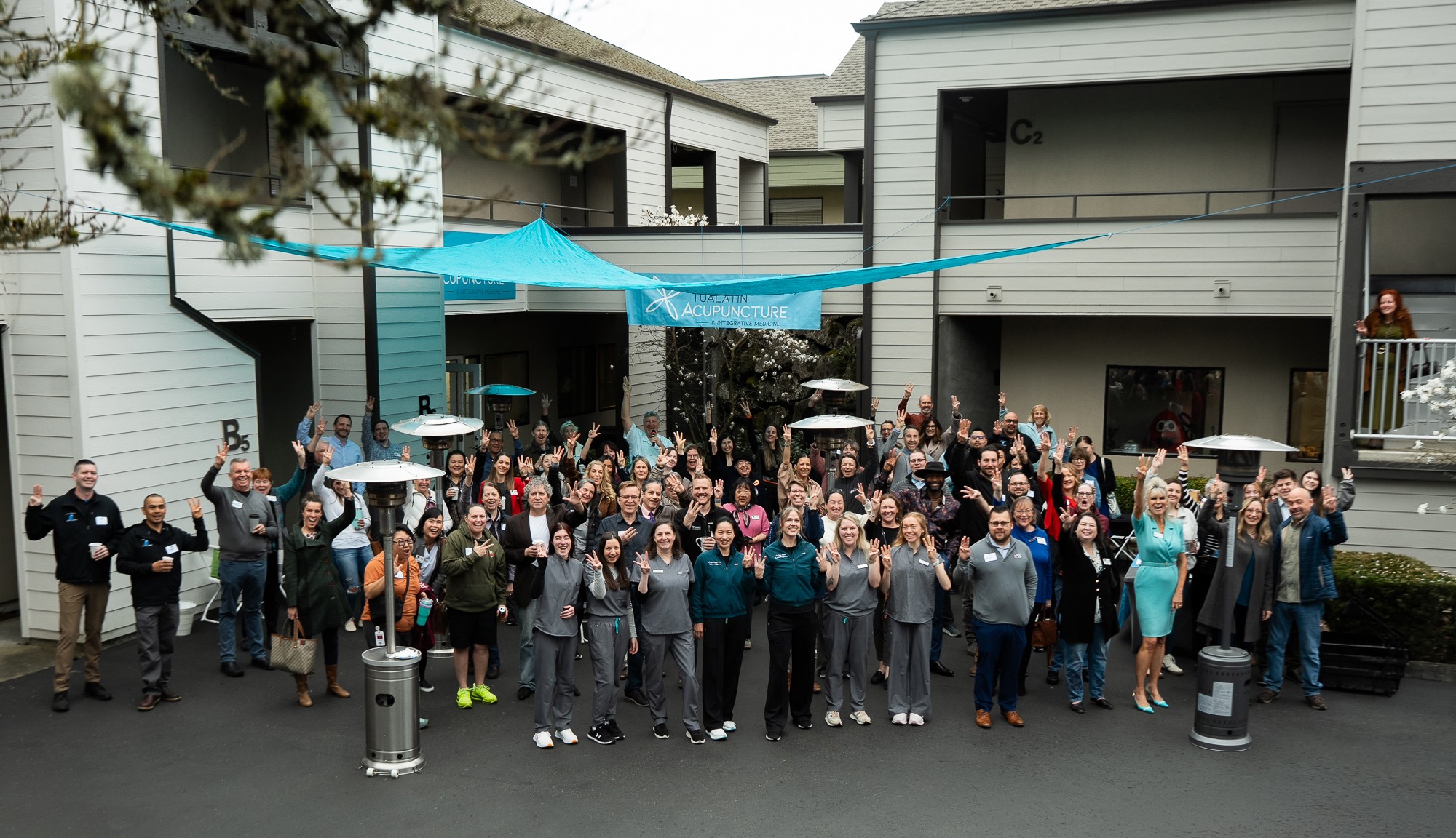 Large group of people gathered outdoors in front of buildings, some holding umbrellas, celebrating together.
