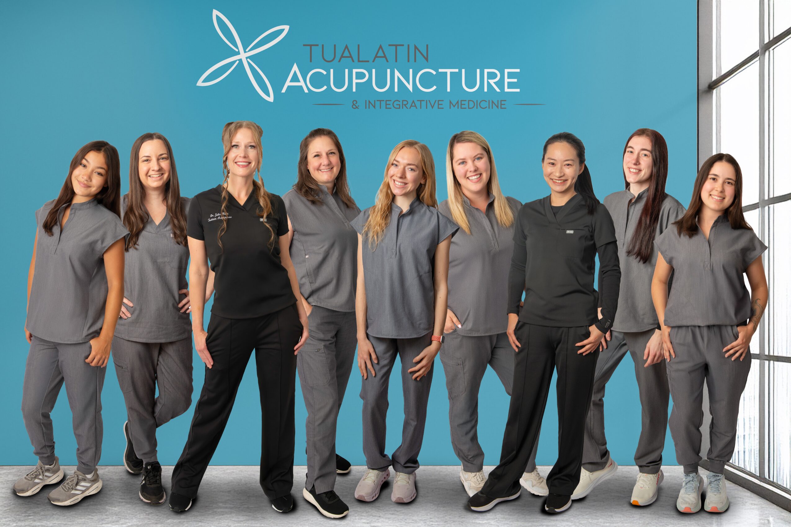 Group of nine women in gray scrubs standing in front of a blue wall with Tualatin Acupuncture logo.