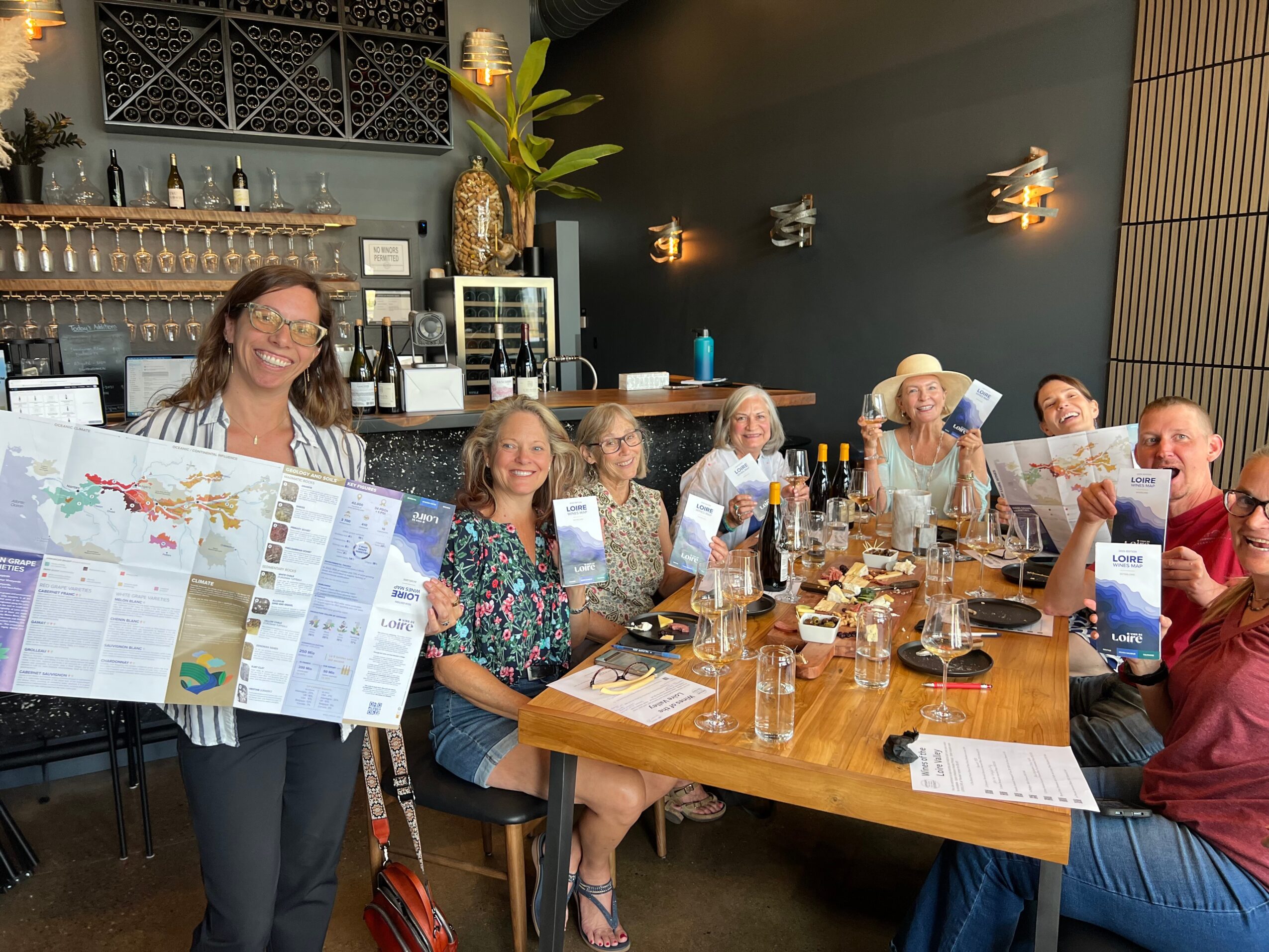 Group of people sitting around a table in a restaurant, with a woman holding a large map, smiling.