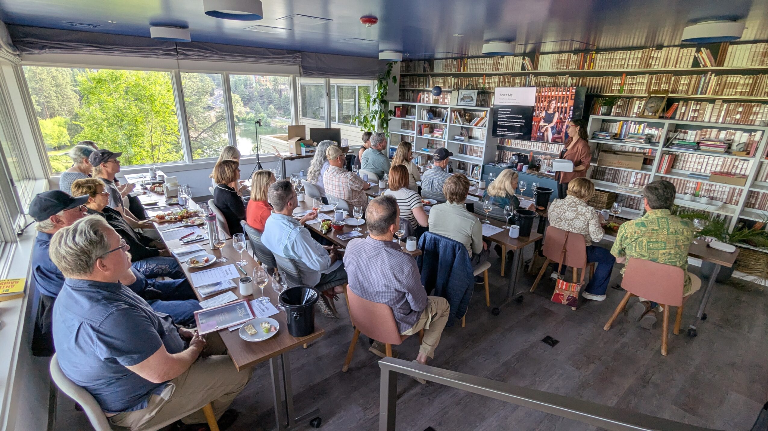 People seated around tables in a room with large windows and bookshelves, attending a presentation or meeting.
