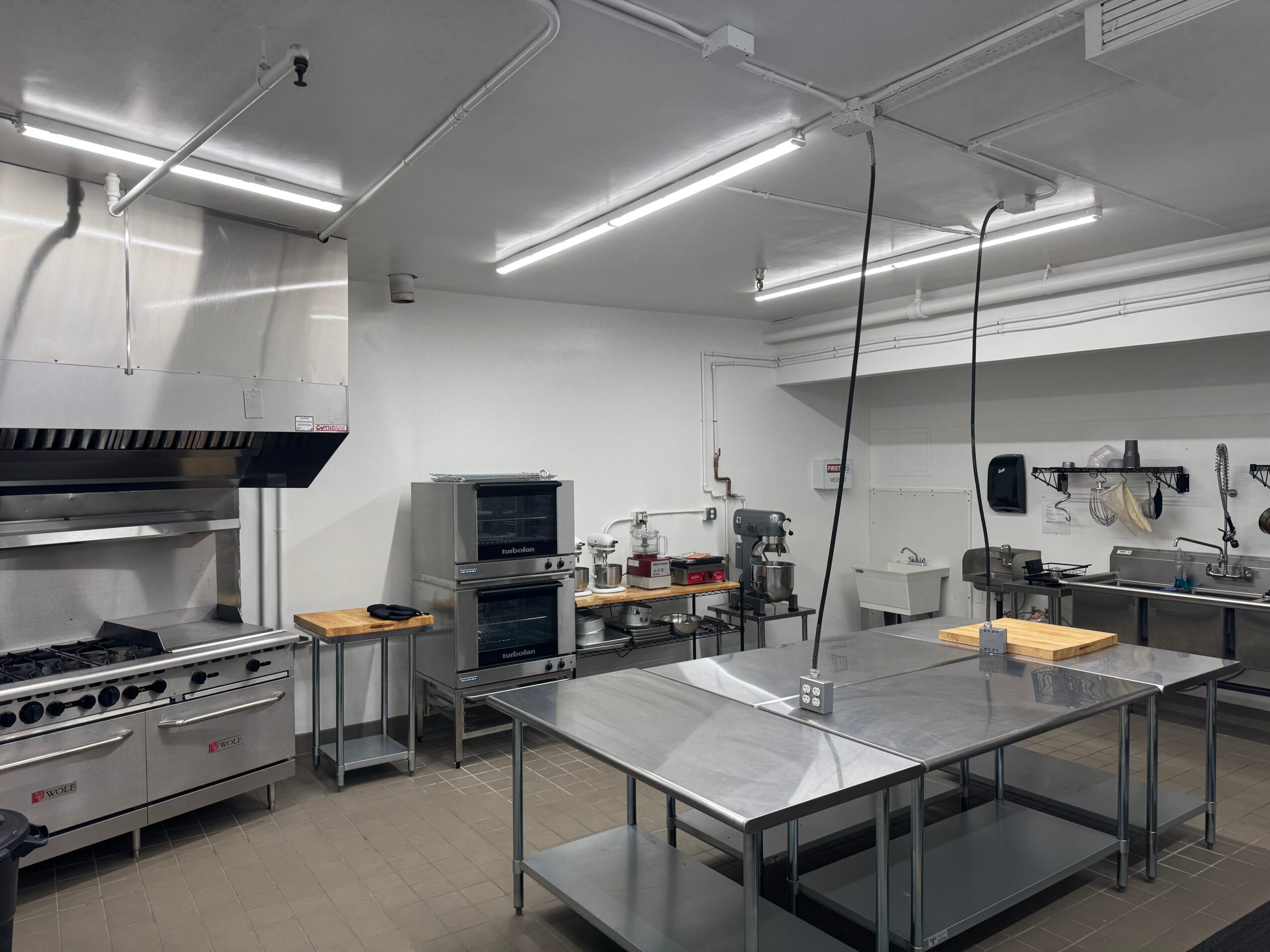 Commercial kitchen with stainless steel tables, appliances, and overhead lighting, viewed from the side with a table in the center.
