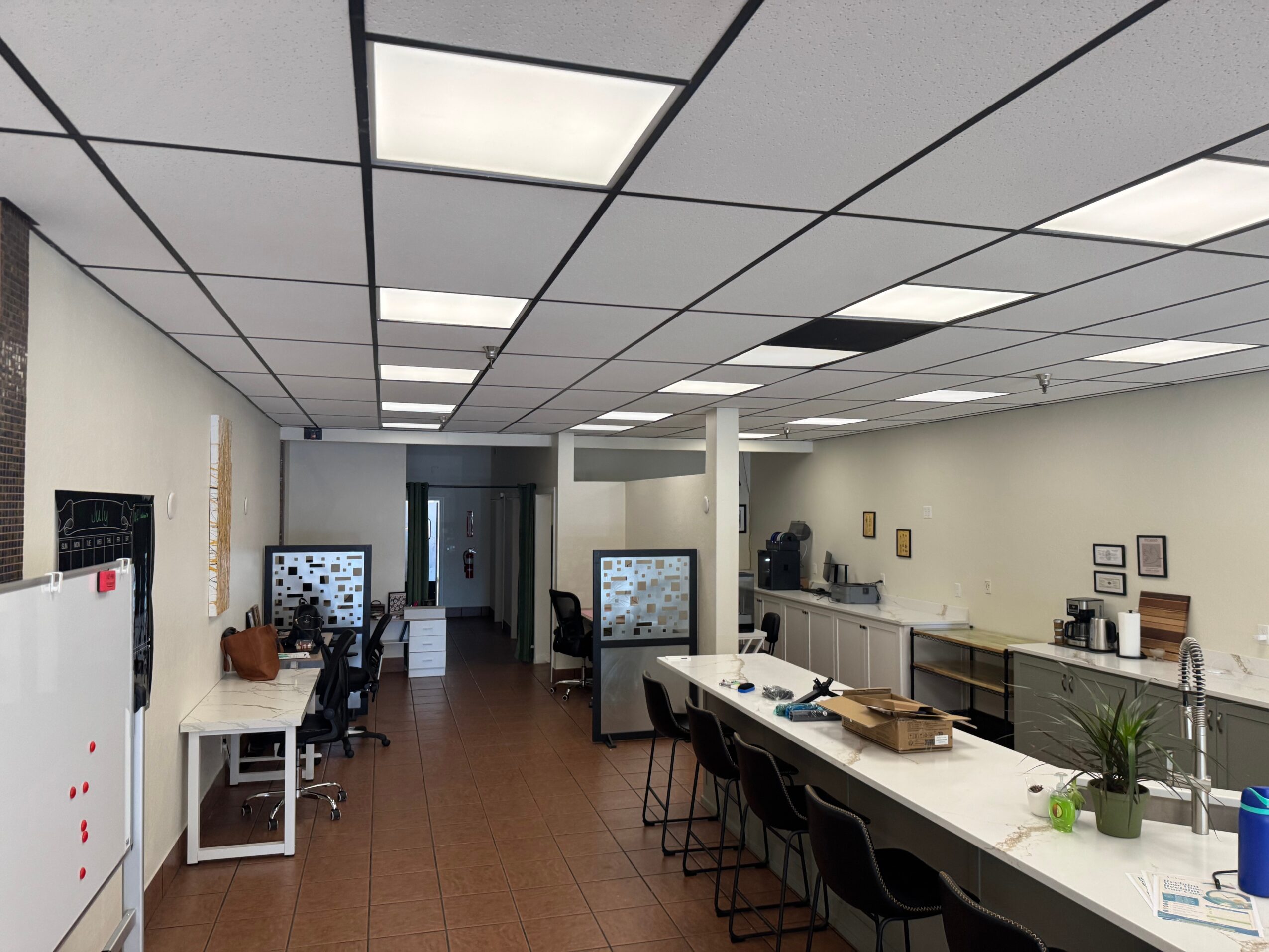 Office space with desks, chairs, and a kitchen area, ceiling with rectangular lights, and a whiteboard on the wall.