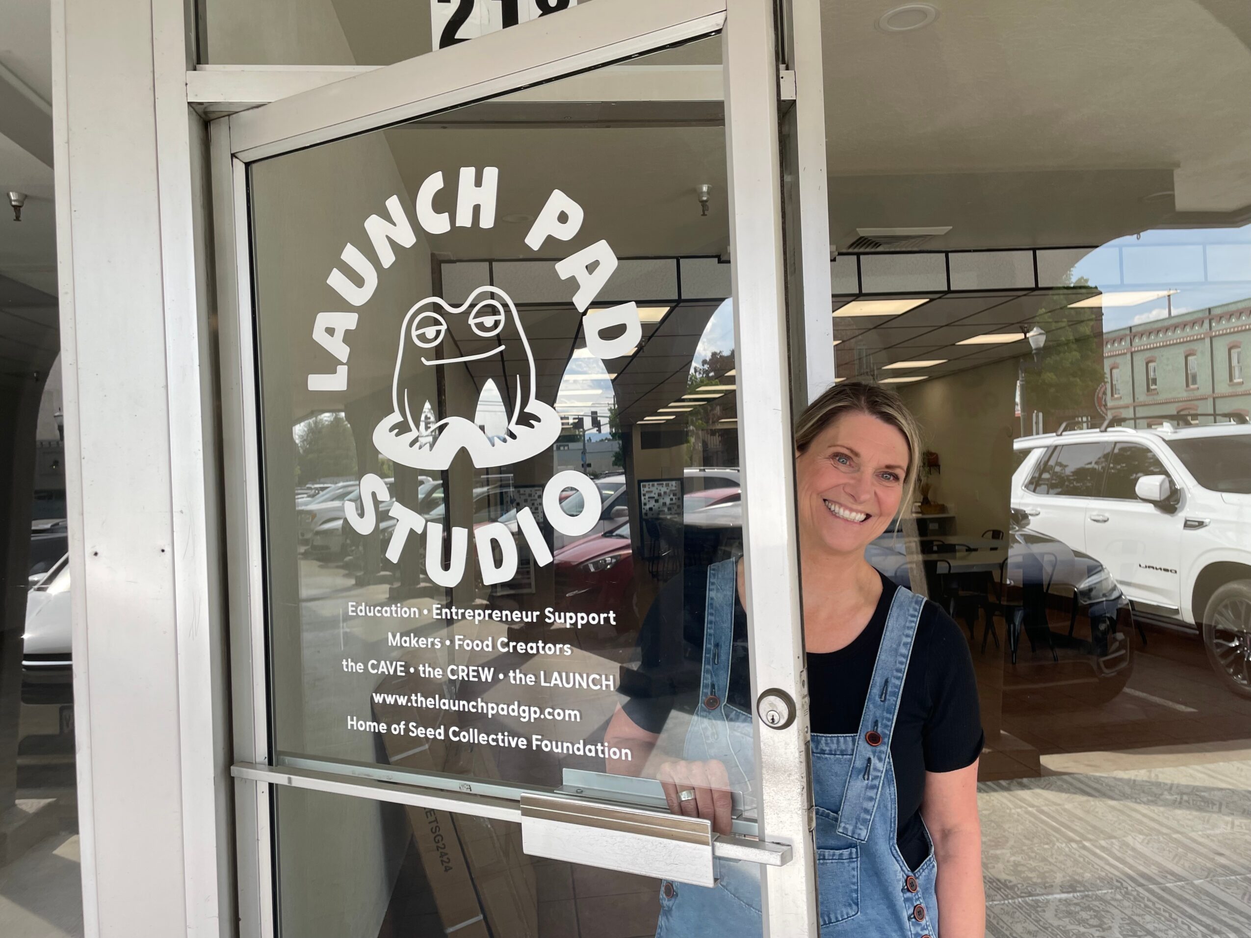 Woman smiling outside a glass door with a logo for Launch Pad Studio, featuring a frog and text, in a sunny environment.
