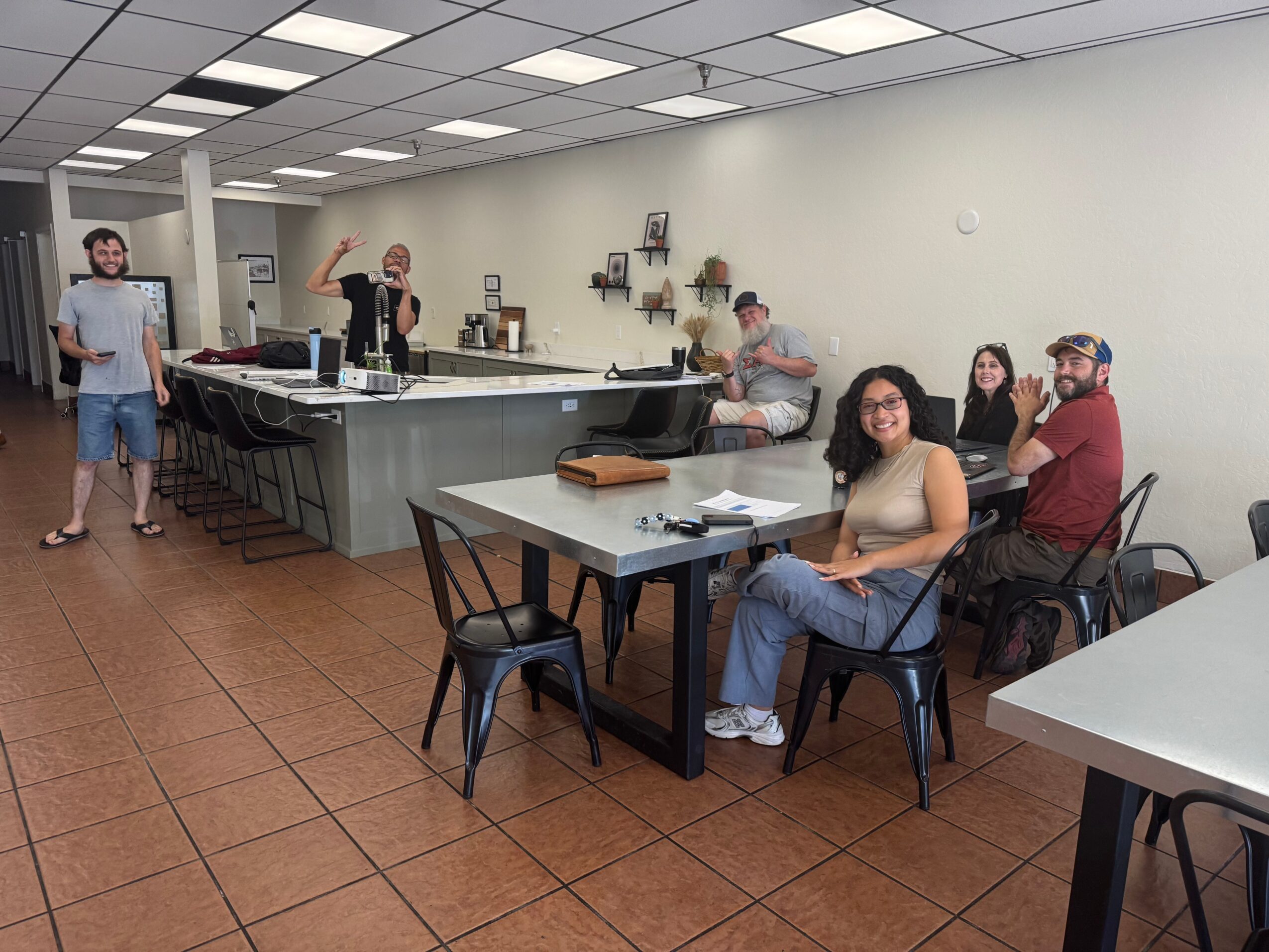 Group of people in a room with tables and chairs, some sitting and some standing, with a counter in the background.
