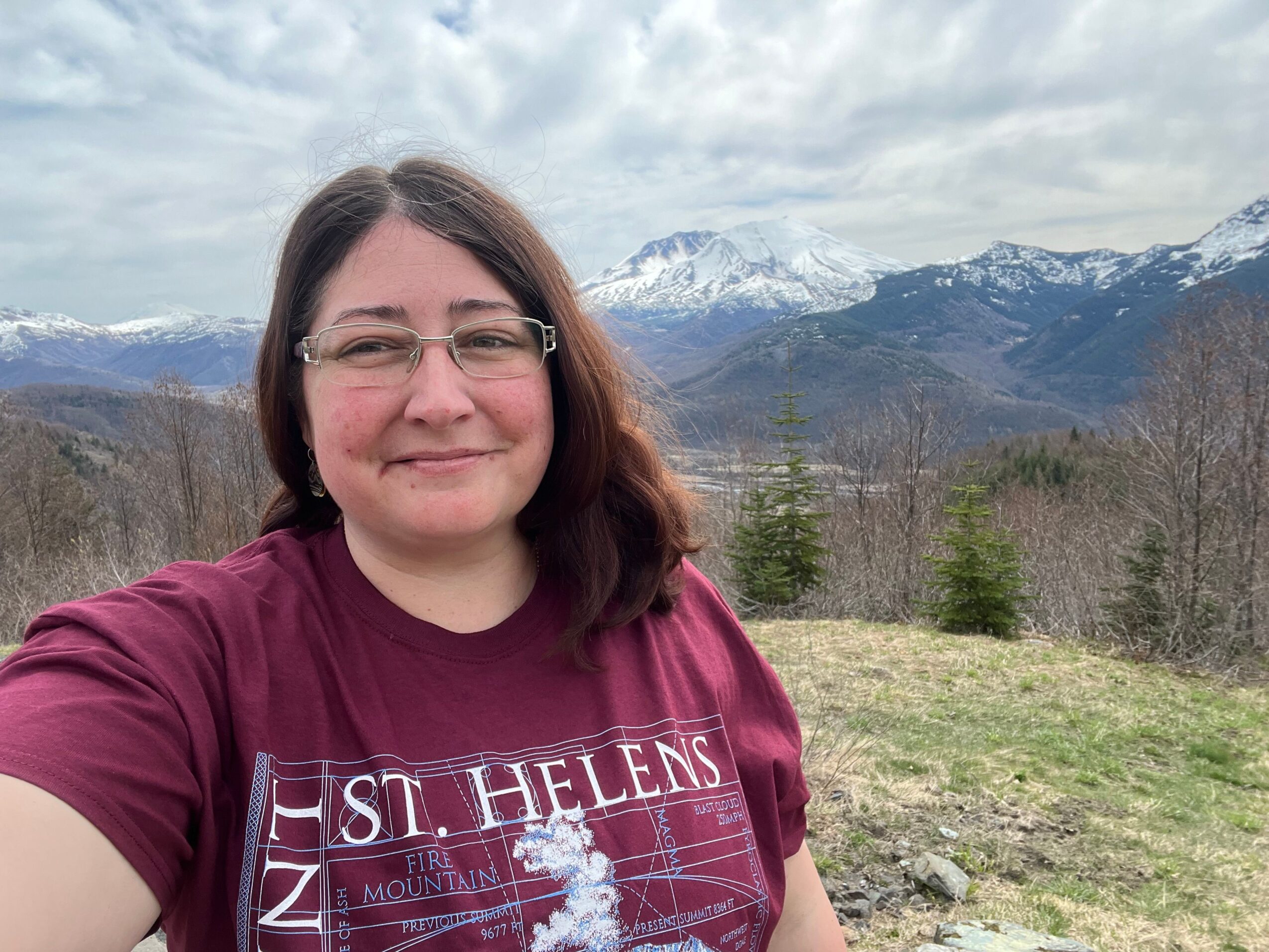 Person with glasses and brown hair outdoors near a body of water, sky with clouds in background.