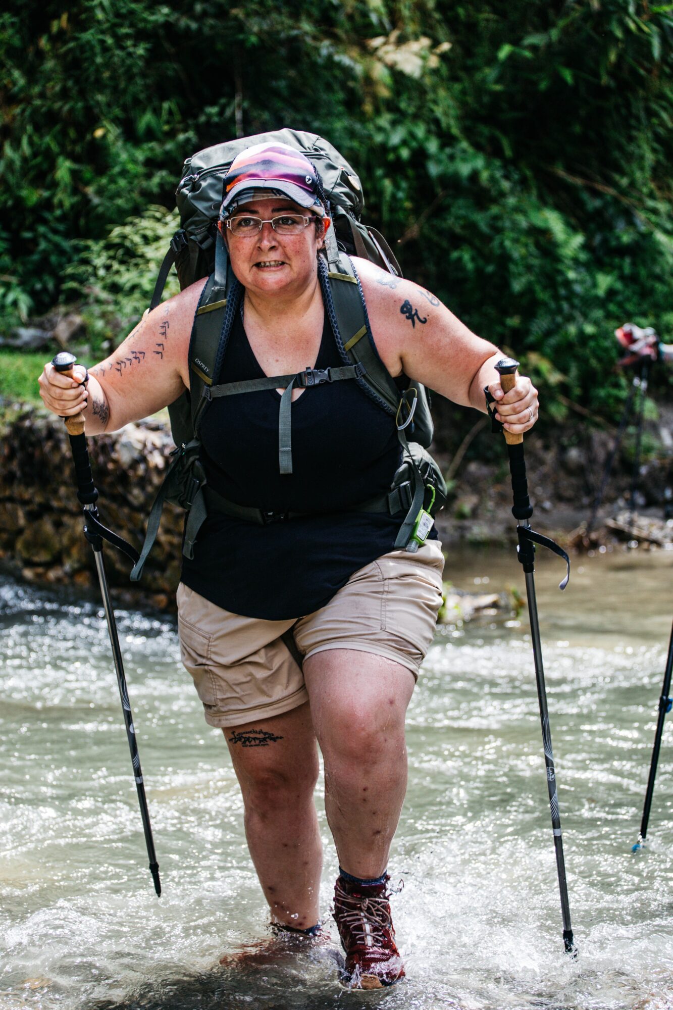 Person hiking through water with a backpack and trekking poles, smiling, surrounded by greenery.