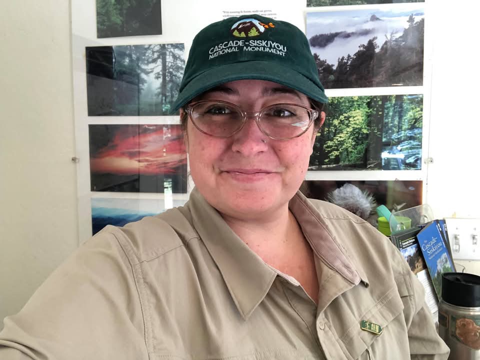 Person wearing glasses, a green cap, and a beige uniform, smiling in front of nature photographs.