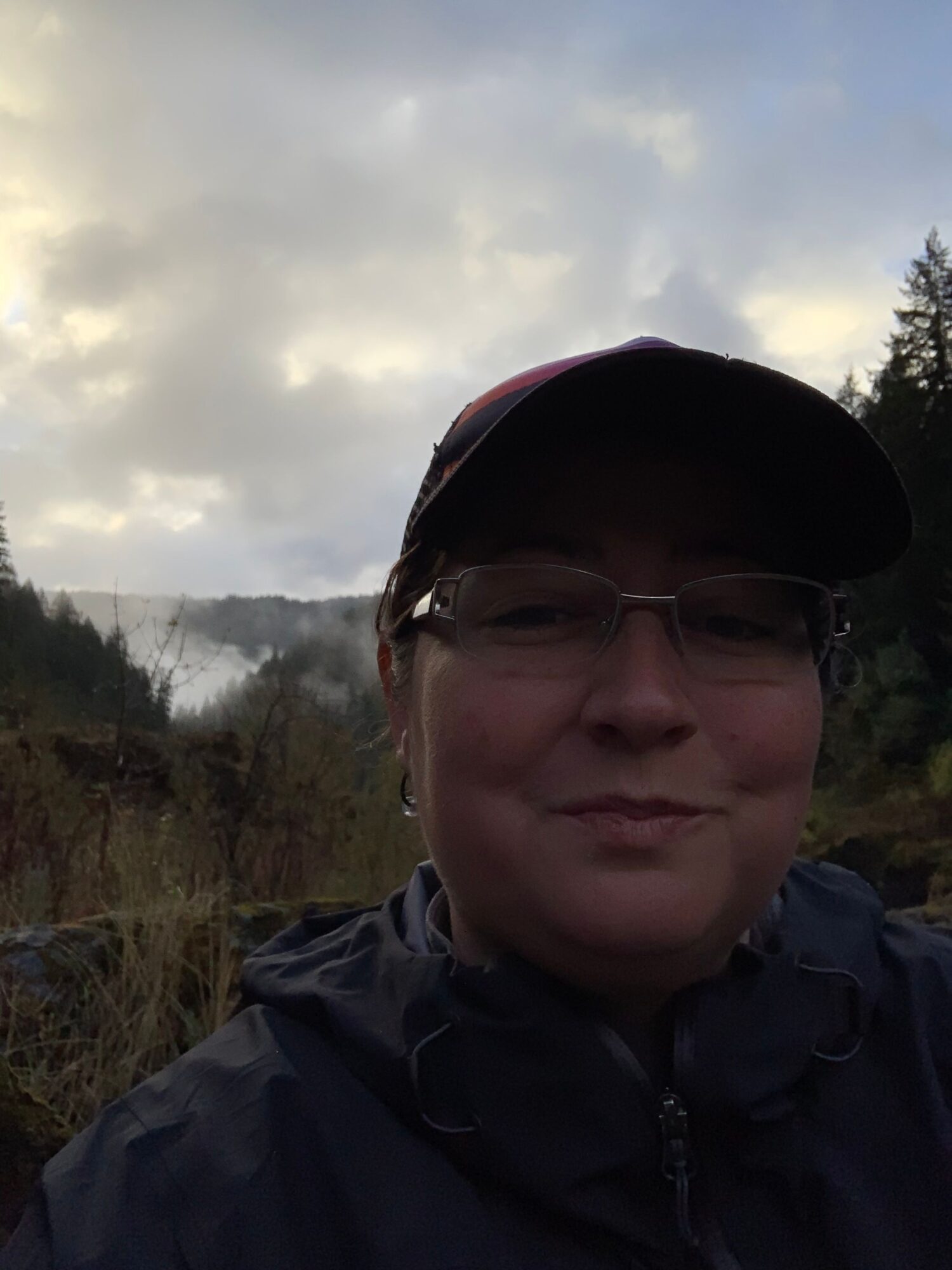 Person wearing glasses and a cap outdoors with cloudy sky and trees in background.