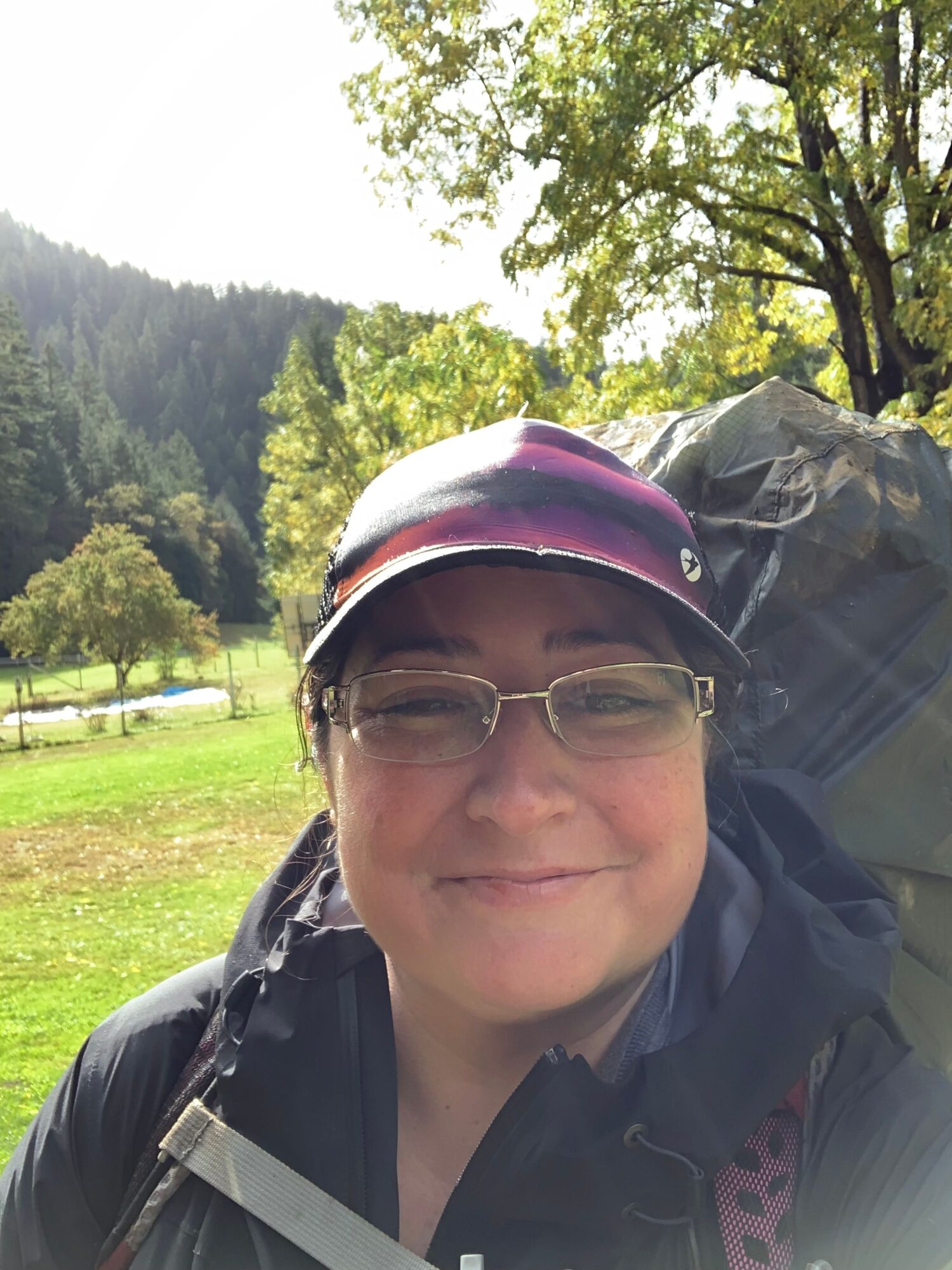 Person wearing glasses and a cap outdoors with trees and grass in the background.