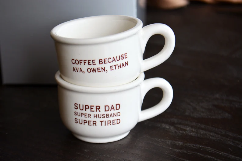 Two stacked white coffee cups with red text on a dark wooden surface.