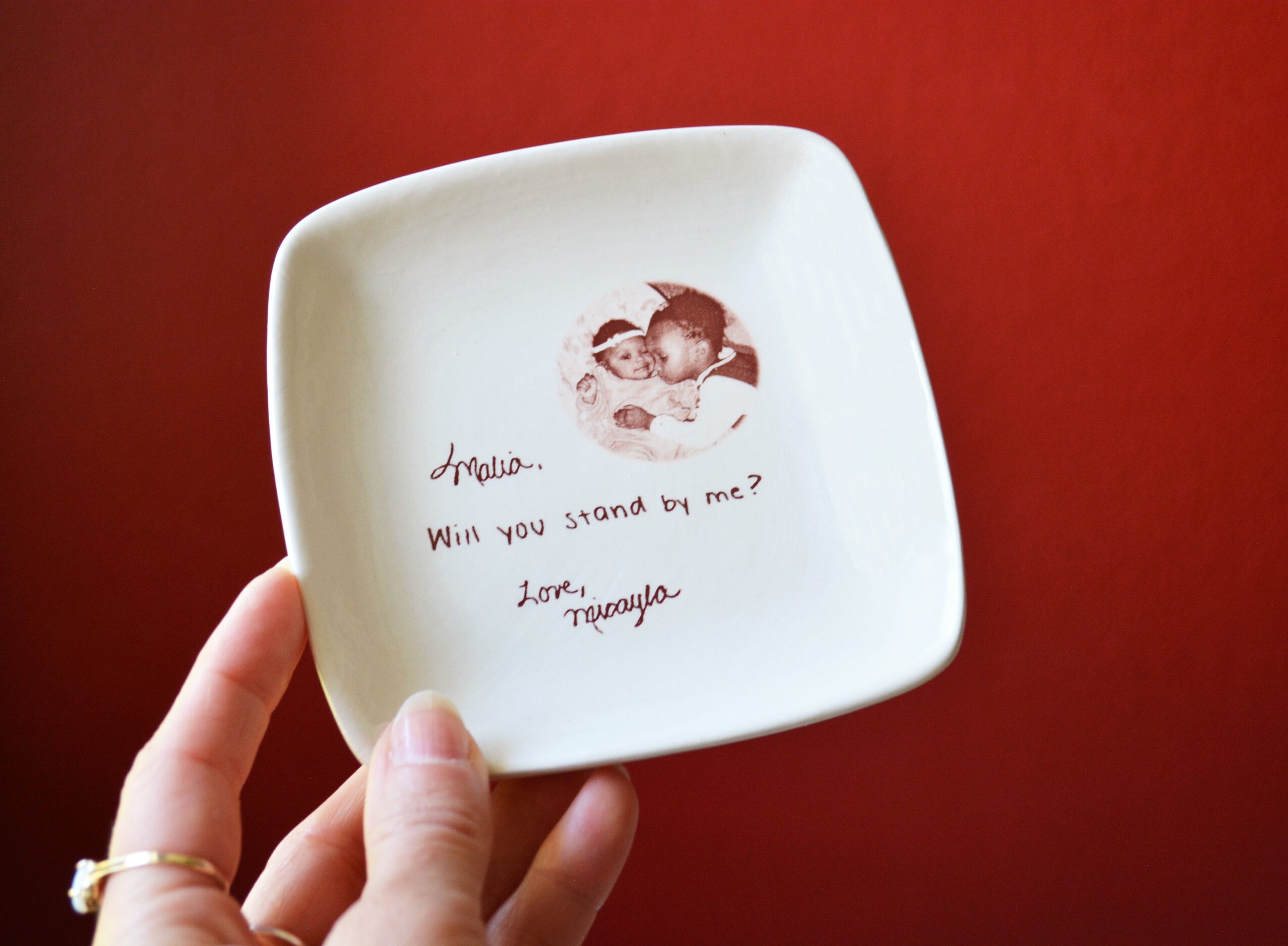 Hand holding a white dish with a photo and handwritten message, against a red background.