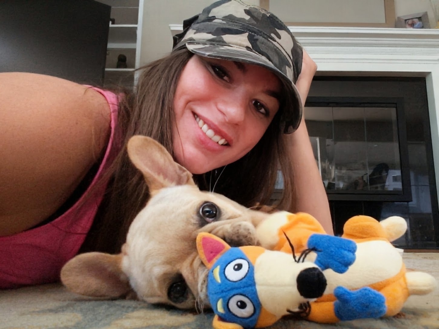 Woman smiling with a puppy holding a colorful plush toy, sitting in front of a fireplace.