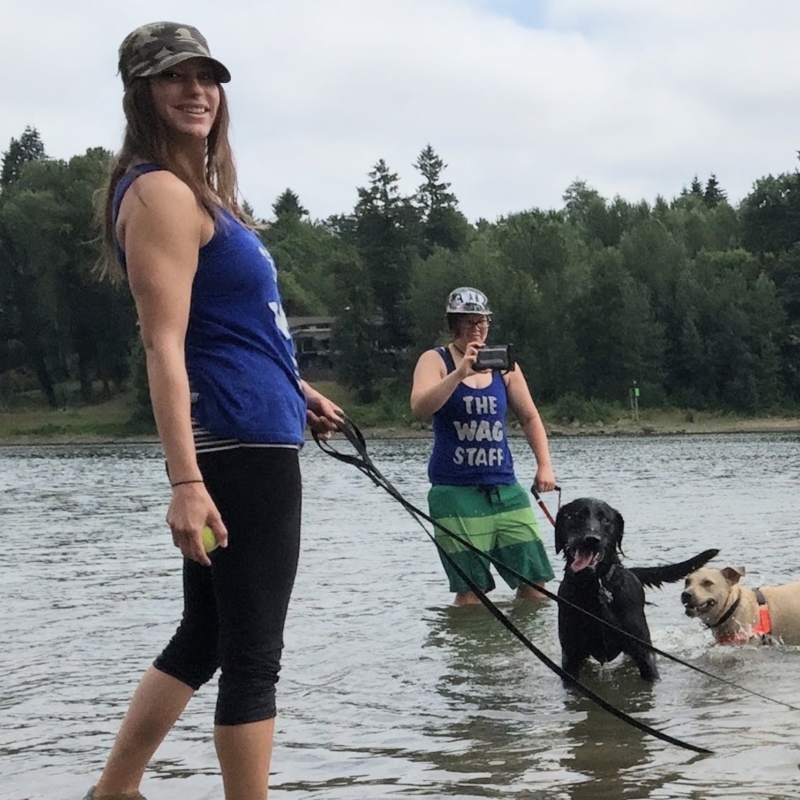 Two people and two dogs standing in a lake with trees in the background, one person taking a photo, one holding a leash.