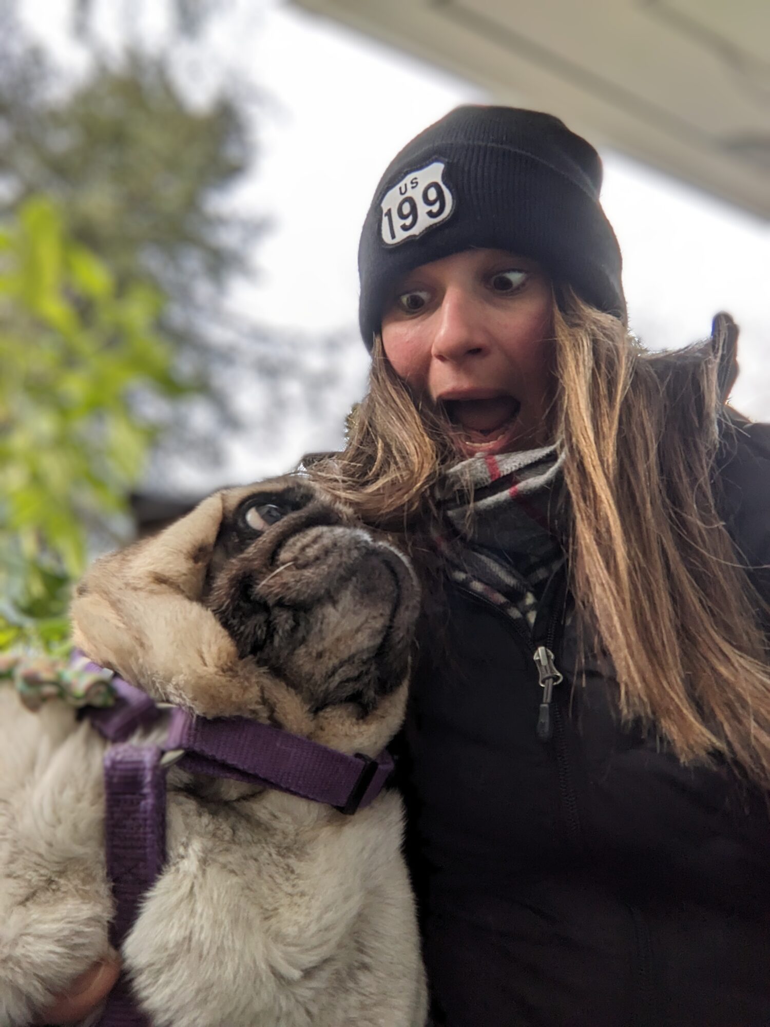 Woman with long hair wearing a black beanie and jacket, surprised expression, with a pug dog wearing a purple harness outdoors.