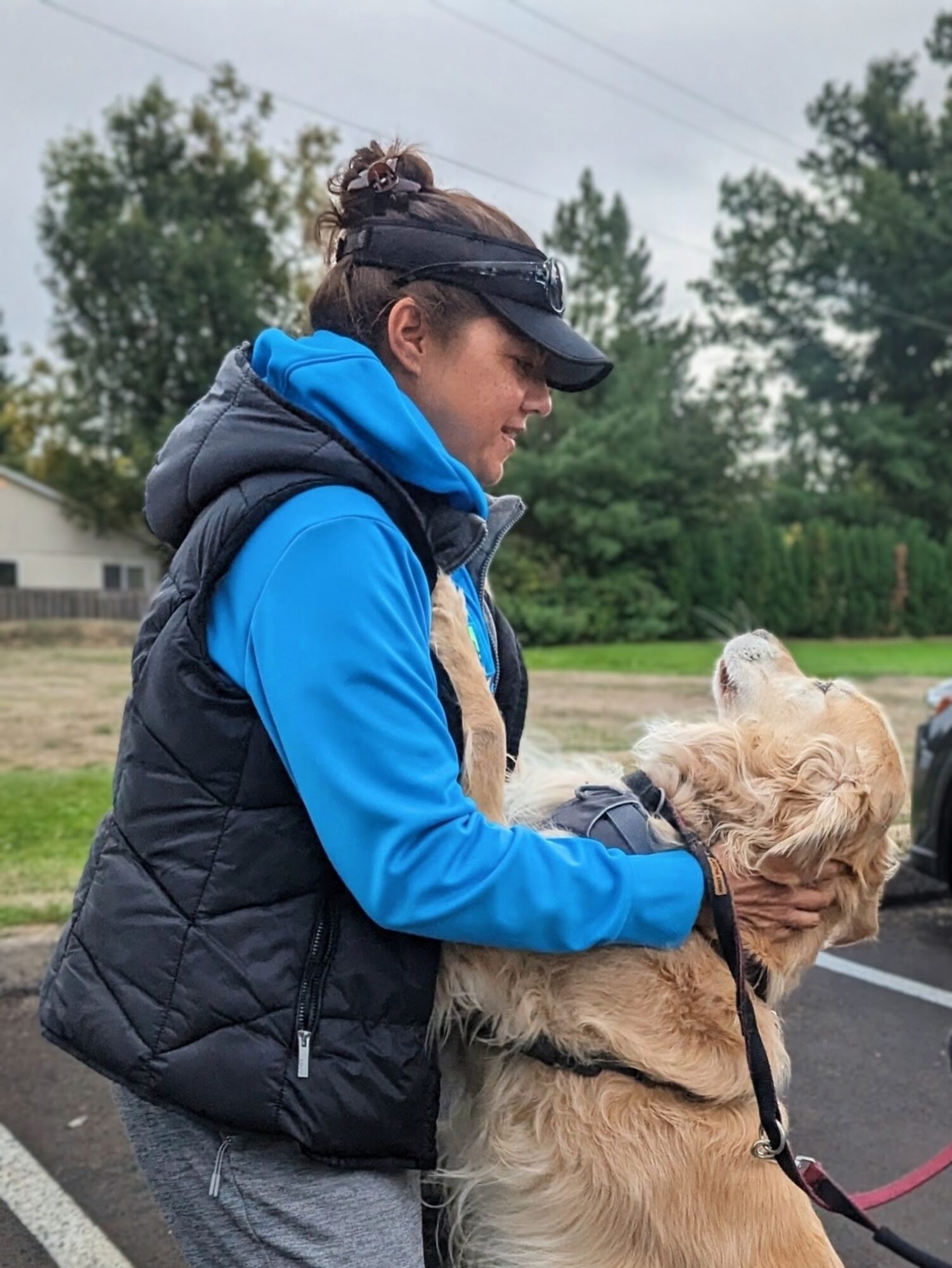 Woman in blue jacket and black vest holding a large dog outdoors, trees and a road in background.