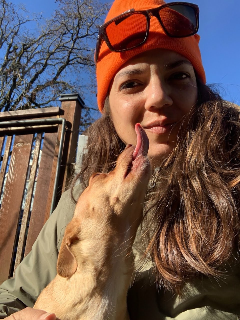 Young woman with long hair wearing an orange beanie and sunglasses, smiling outdoors with a dog touching her face.