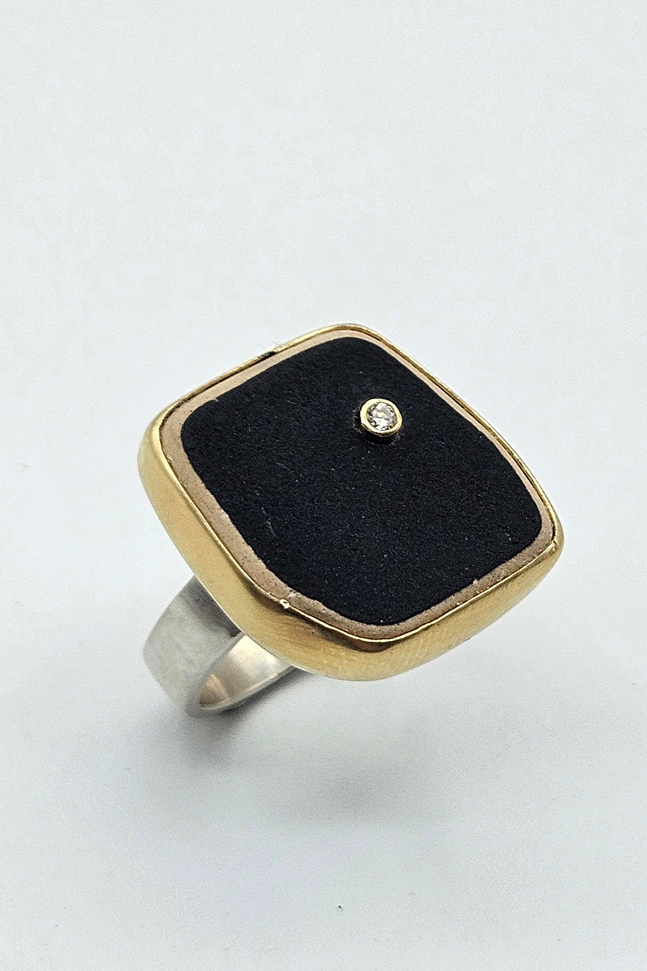 Ring with a square face, black background, small central gemstone, gold border, and silver band, on a plain background.