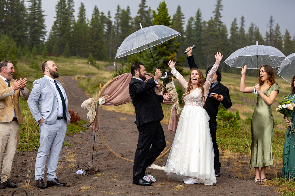Group of people celebrating outdoors in a forested area, some holding umbrellas, with a woman in a white dress and others clapping.