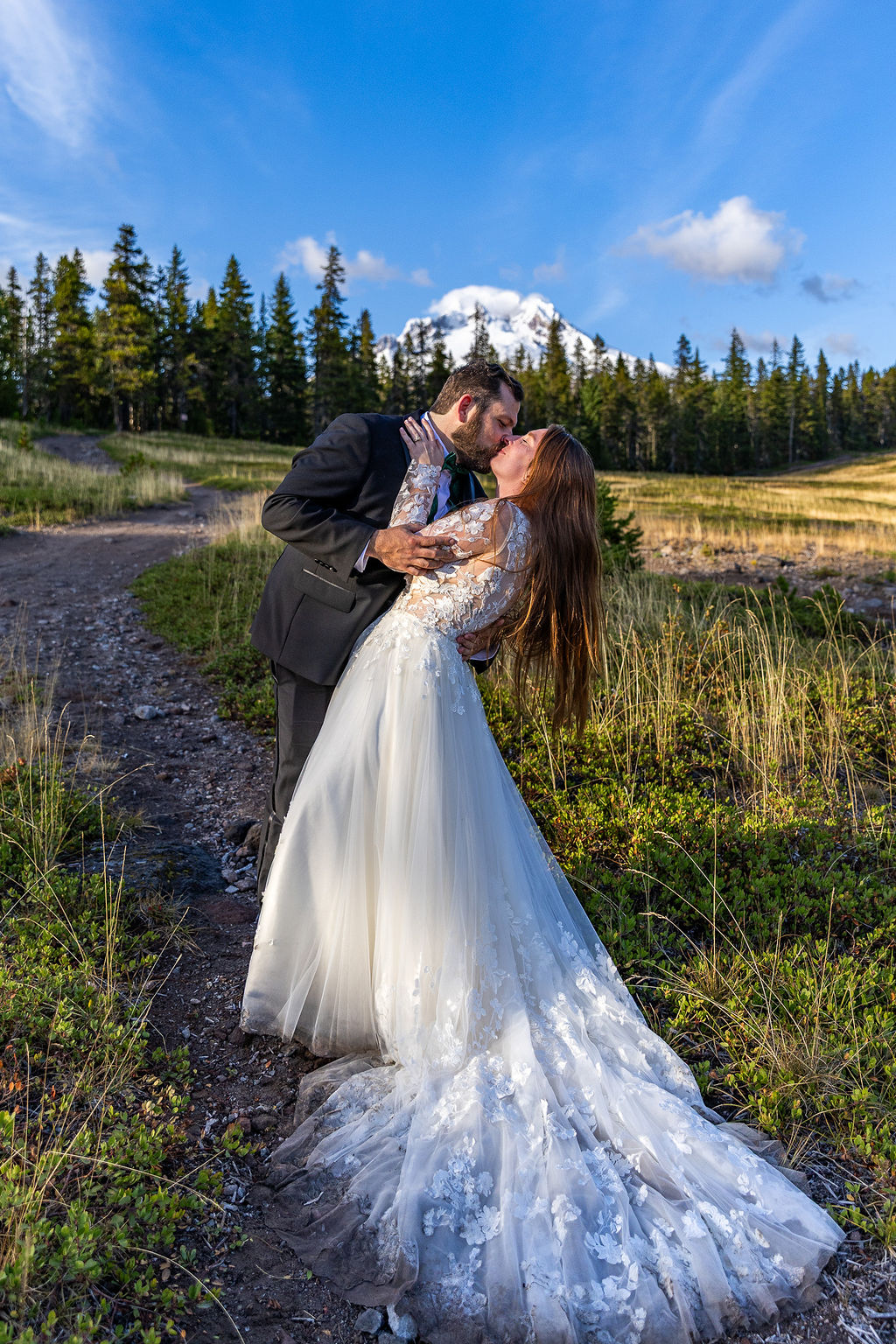 A couple in wedding attire kissing outdoors on a grassy path with trees and a snow-capped mountain in the background.