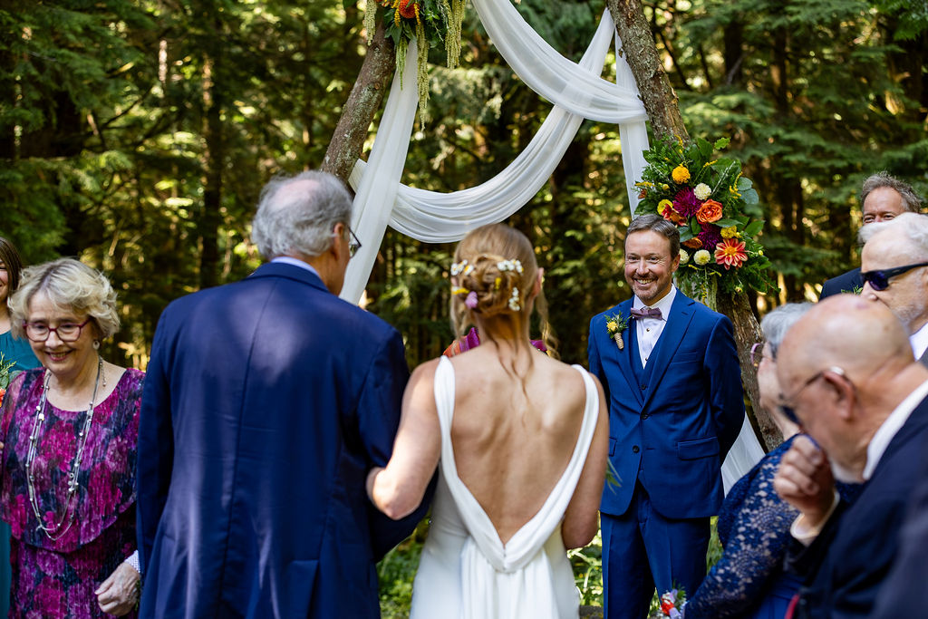 People gathered outdoors for a wedding ceremony, with a woman in a white dress and a man in a blue suit smiling, surrounded by guests and decorated arch.