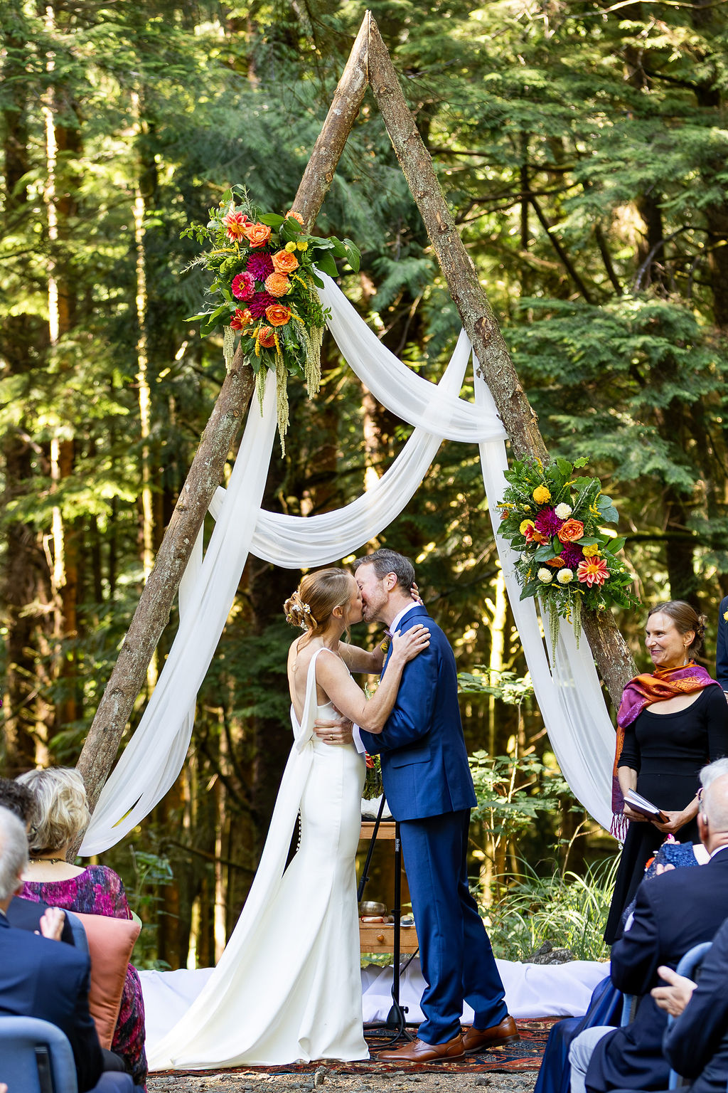 Couple kissing under a wooden arch decorated with flowers and white fabric in a forest setting.