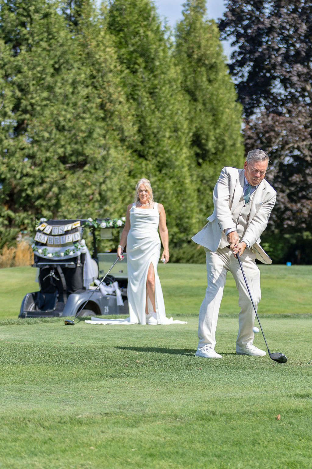 A man in a light suit is playing golf on a green, with a woman in a white dress watching nearby, trees in background.