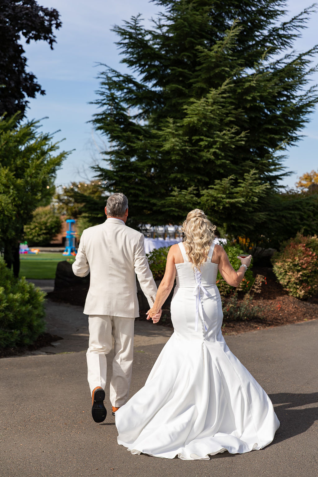 A bride and groom walk hand in hand outdoors, surrounded by trees and greenery, with the bride in a white wedding gown.
