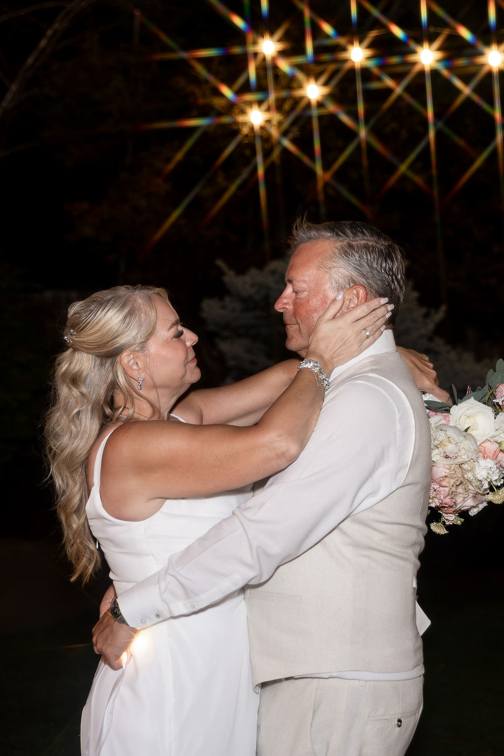 A couple dancing outdoors at night, with string lights above and flowers nearby, gazing at each other.