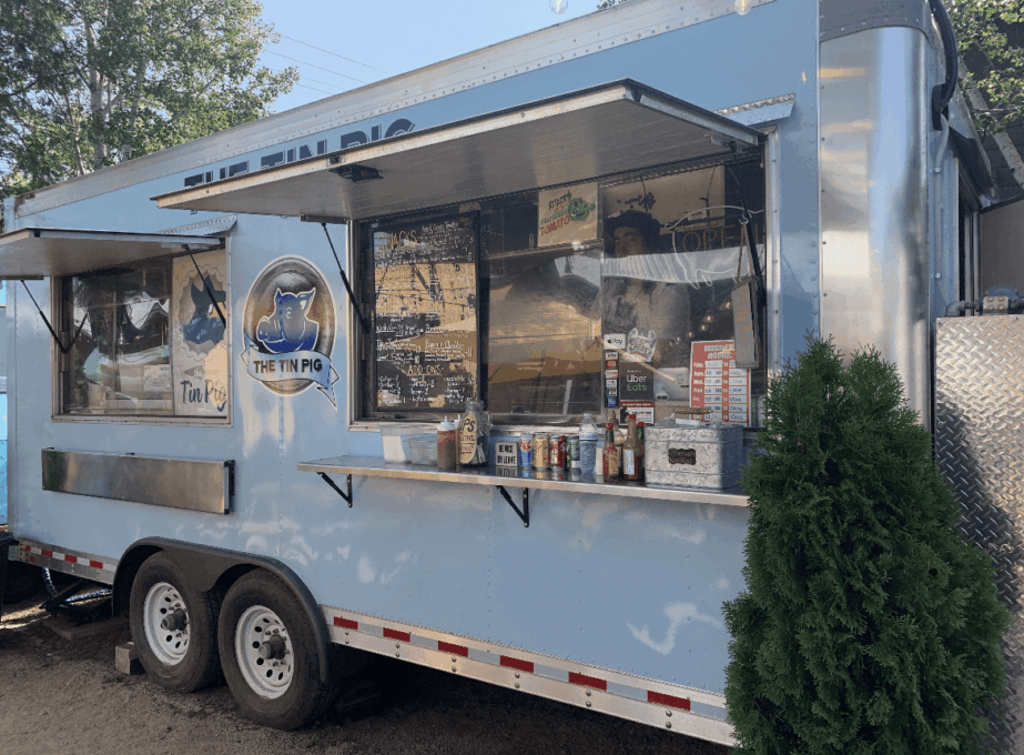 Light blue food truck with large window, parked outdoors with trees and a bush nearby.