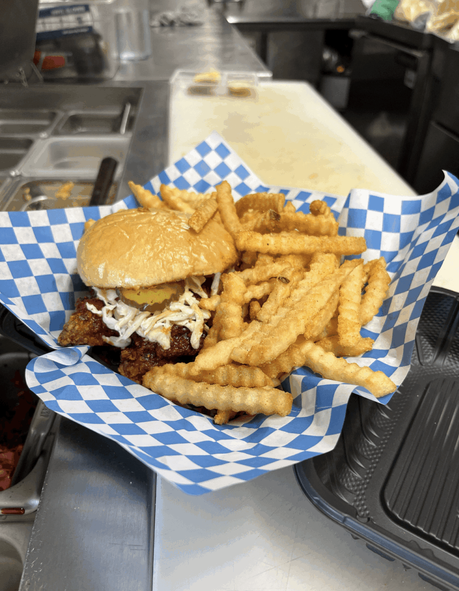 Sandwich with fries on blue checkered paper, placed on a metal counter in a kitchen setting.