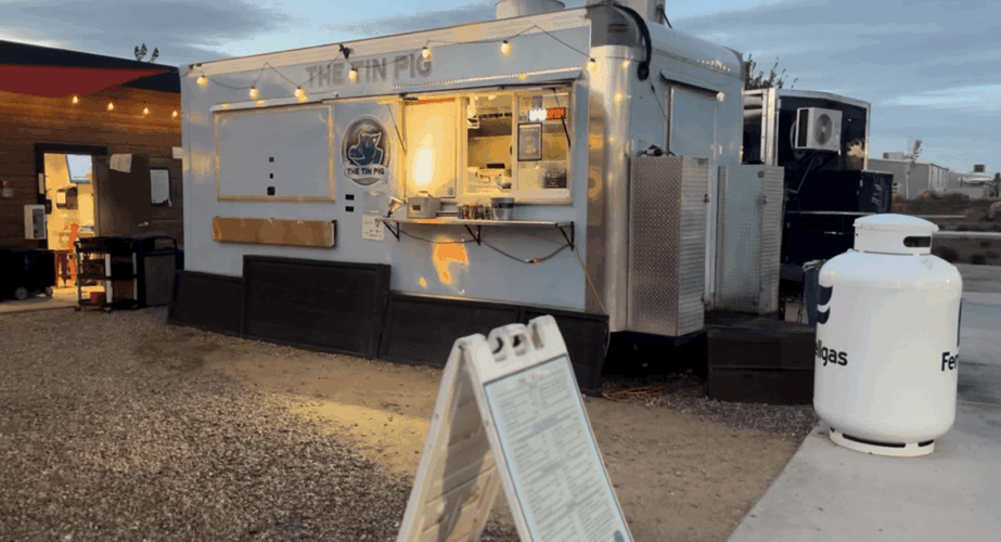 Food truck with a serving window, parked outdoors with a barrel nearby, under a clear sky.