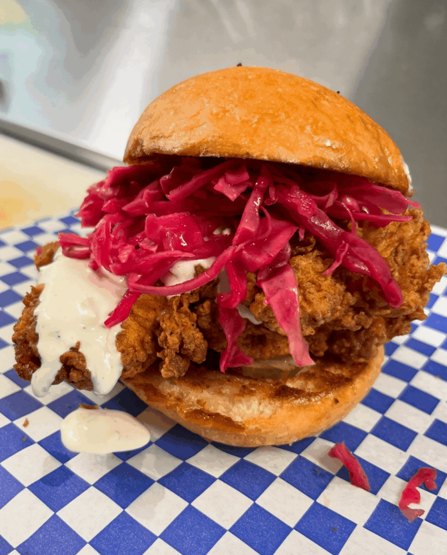 Fried chicken sandwich with pickled red onions and sauce on a bun, served on blue checkered paper.
